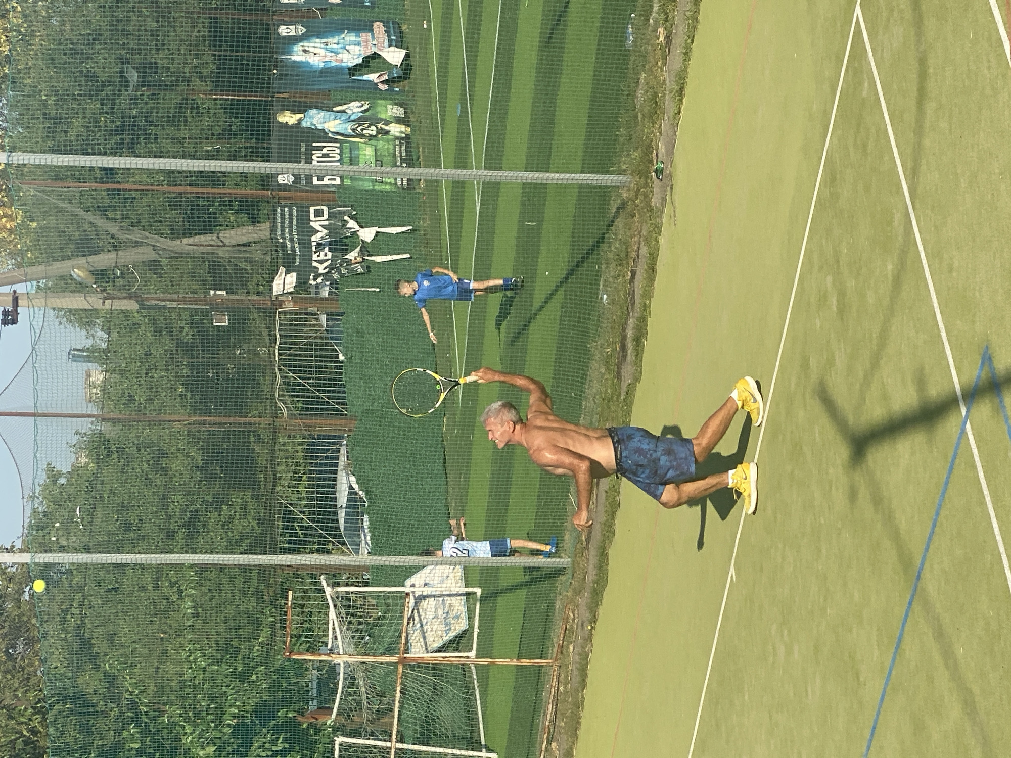 The image depicts an outdoor tennis court with a man in the foreground mid-serve, his body angled upwards as he prepares to strike a tennis ball suspended in the air. He is shirtless, wearing dark patterned shorts and bright yellow tennis shoes. In the middle ground, a young boy in a blue shirt and shorts is visible on the court, also appearing to be engaged in a game. Further in the background, another young boy in a blue shirt with the number '42' on it stands near a soccer goal. The court itself is green with white and blue court lines. A chain-link fence encloses the court, with trees and buildings visible beyond. Posters are affixed to the fence in the background, some of which are partially torn. The scene appears to be set in a sports complex, possibly in the city of Krasnodar, Russia, given the presence of Cyrillic text on the posters in the background. The lighting suggests it is a sunny day.