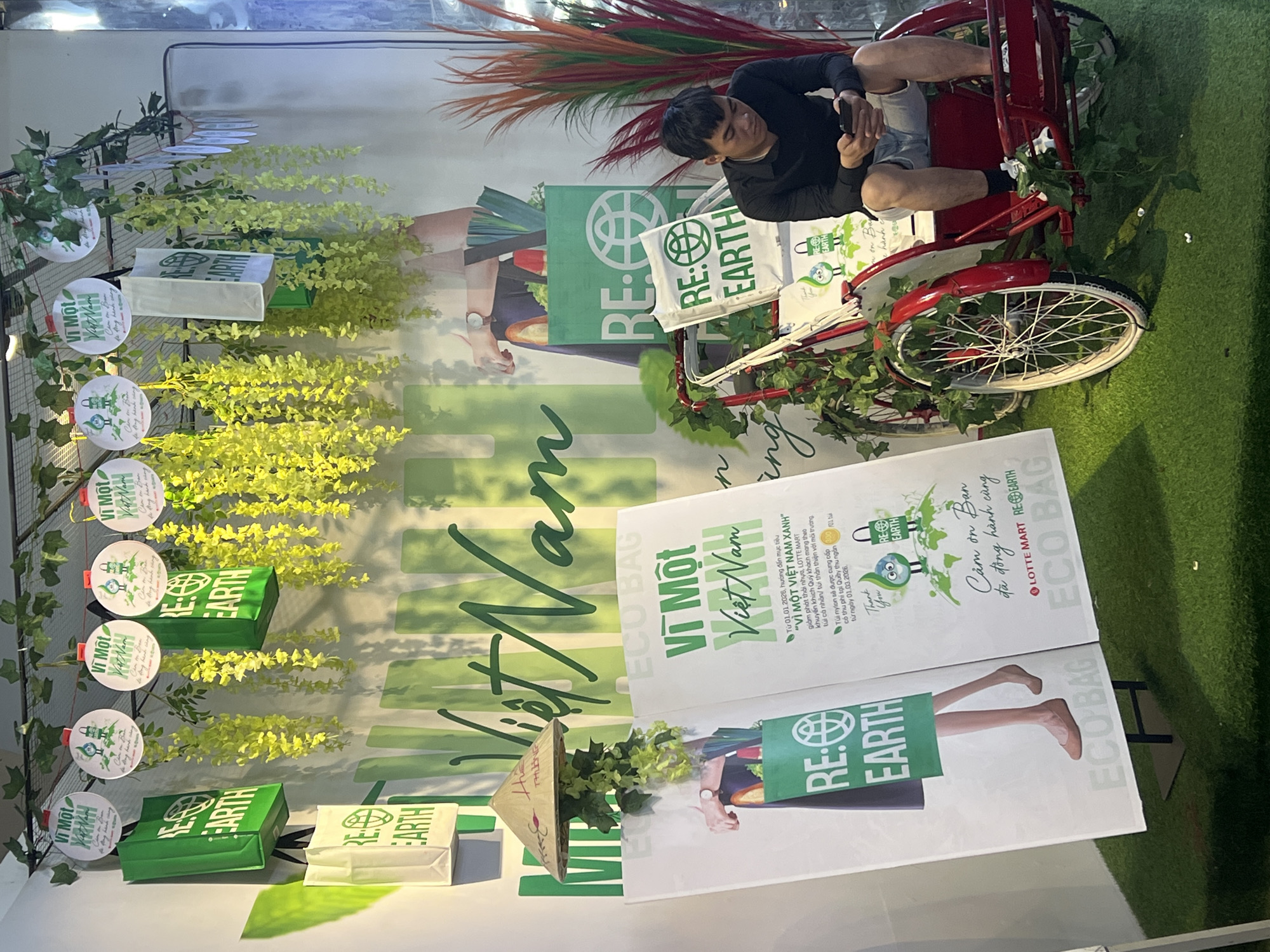 The image shows a promotional display in Đà Nẵng, Vietnam, likely inside a store or at an event. The display promotes an eco-friendly initiative called "RE: EARTH" with the slogan "For a Green Vietnam". Green and yellow floral arrangements and signage with Vietnamese text dominate the display. Tote bags with the "RE: EARTH" logo are visible. A man sits on a red tricycle, partially obscured by the display, and appears to be looking at his phone. The ground is covered with artificial green grass.