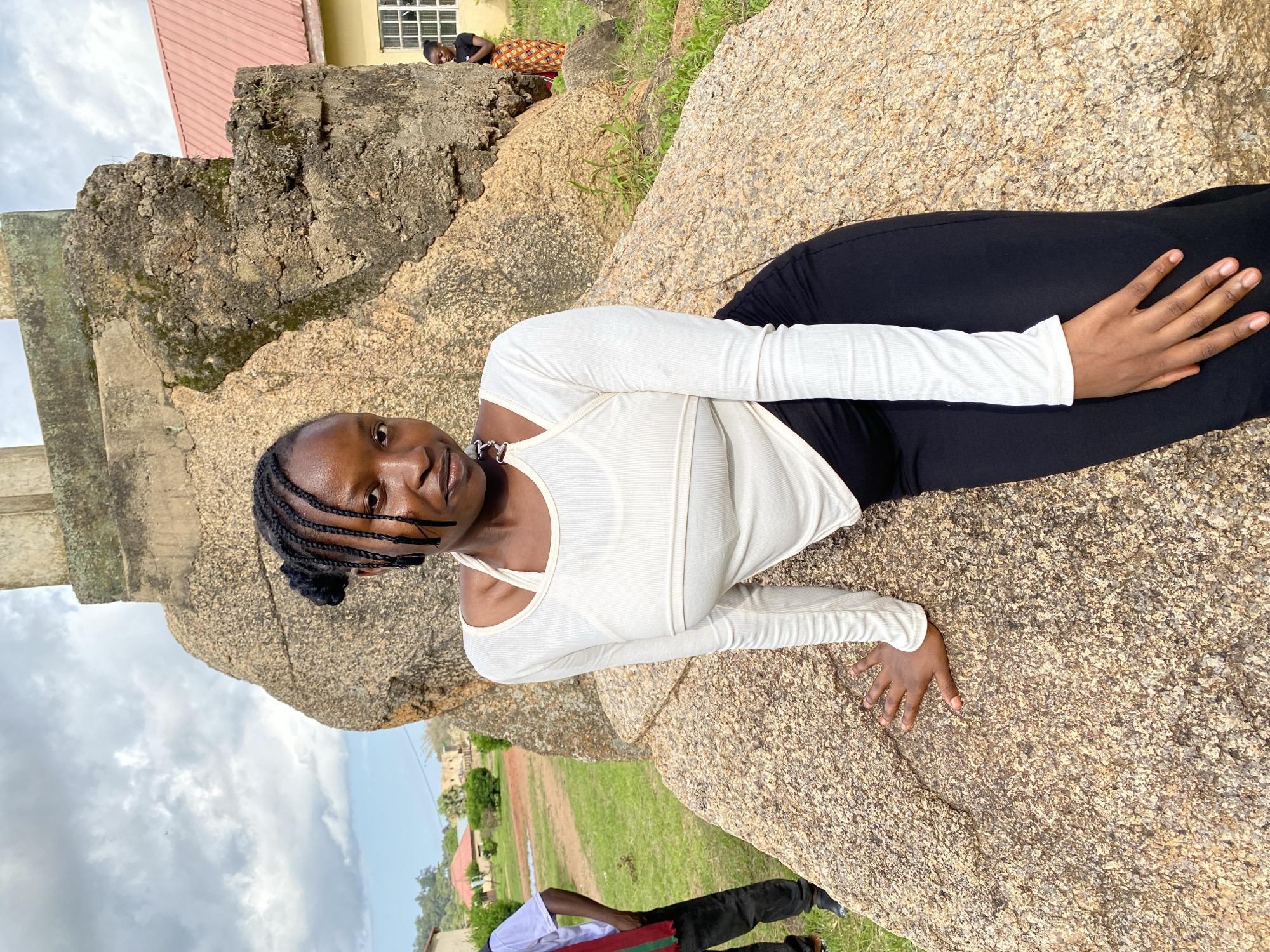 A young Black woman with braided hair is leaning on a large boulder outdoors. She is wearing a white long-sleeved crop top and black pants. She is looking at the camera with a slight smile. In the background, there are buildings and trees. One building appears to be a school or administrative building with a red roof. Another person, wearing a white shirt and black pants, is standing to the left of the main subject. Further in the background, on the right, another young Black woman is visible near the building. The sky is cloudy. The environment appears to be a public space or park within a city. The scene is likely located in the city of Wang, Nigeria, given the context.