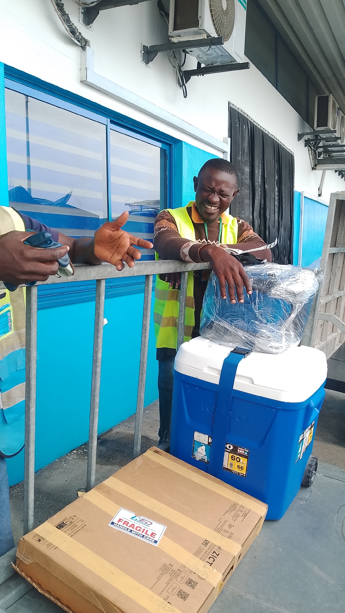 In Orerokpe, Nigeria, a man wearing a high-visibility vest laughs heartily as he leans on a metal railing. He is standing next to a blue and white Igloo cooler, which has a blue strap across its lid. On top of the cooler, there is a package wrapped in clear plastic. In the foreground, a brown cardboard box with "FRAGILE HANDLE WITH CARE" printed on a white sticker rests on the ground. The background features a building with bright blue siding and windows, and an air conditioning unit mounted on the wall. Another person's hands are visible on the railing to the left, with one hand gesturing outwards.