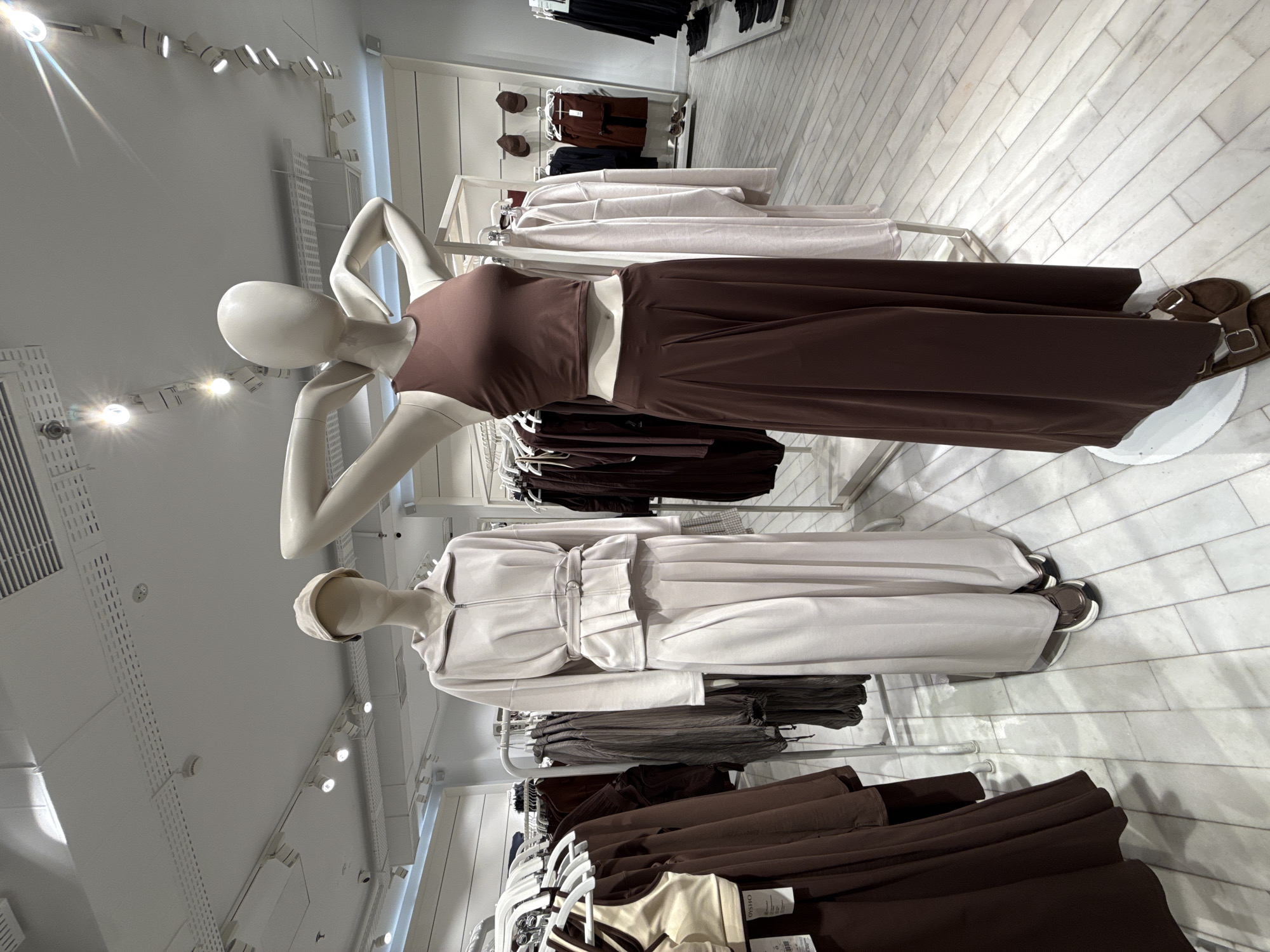 This image captures the interior of a modern clothing store, likely in Kuta, Indonesia, showcasing fashion items on two mannequins and multiple clothing racks. The scene is presented horizontally, with the subjects appearing rotated 90 degrees from their typical upright position.
In the foreground, a white mannequin in a dynamic pose, with its arms raised behind its head, wears a two-piece ensemble. This outfit consists of a form-fitting, sleeveless brown crop top and matching wide-leg, flowing brown trousers. Brown slip-on sandals with buckled straps complete its attire.
Behind and slightly below the first, a second white mannequin, distinguished by a light-colored cap, displays a monochromatic light grey or off-white outfit. This includes a belted jacket with a broad collar and long sleeves, paired with coordinating wide-leg pants. It also wears similar brown slip-on sandals.
Numerous clothing racks filled with garments on white hangers surround the mannequins, featuring items in a palette of browns, blacks, and light greys/off-whites. A small tag partially visible on a lower rack shows the brand name "OYSHO."
The store environment is brightly lit by white track spotlights mounted on the ceiling. The walls are white, and the floor is covered in light-colored, rectangular tiles arranged in a stretcher bond pattern, creating a clean and contemporary aesthetic. The overall impression is that of a minimalist and well-organized retail display focused entirely on the apparel.