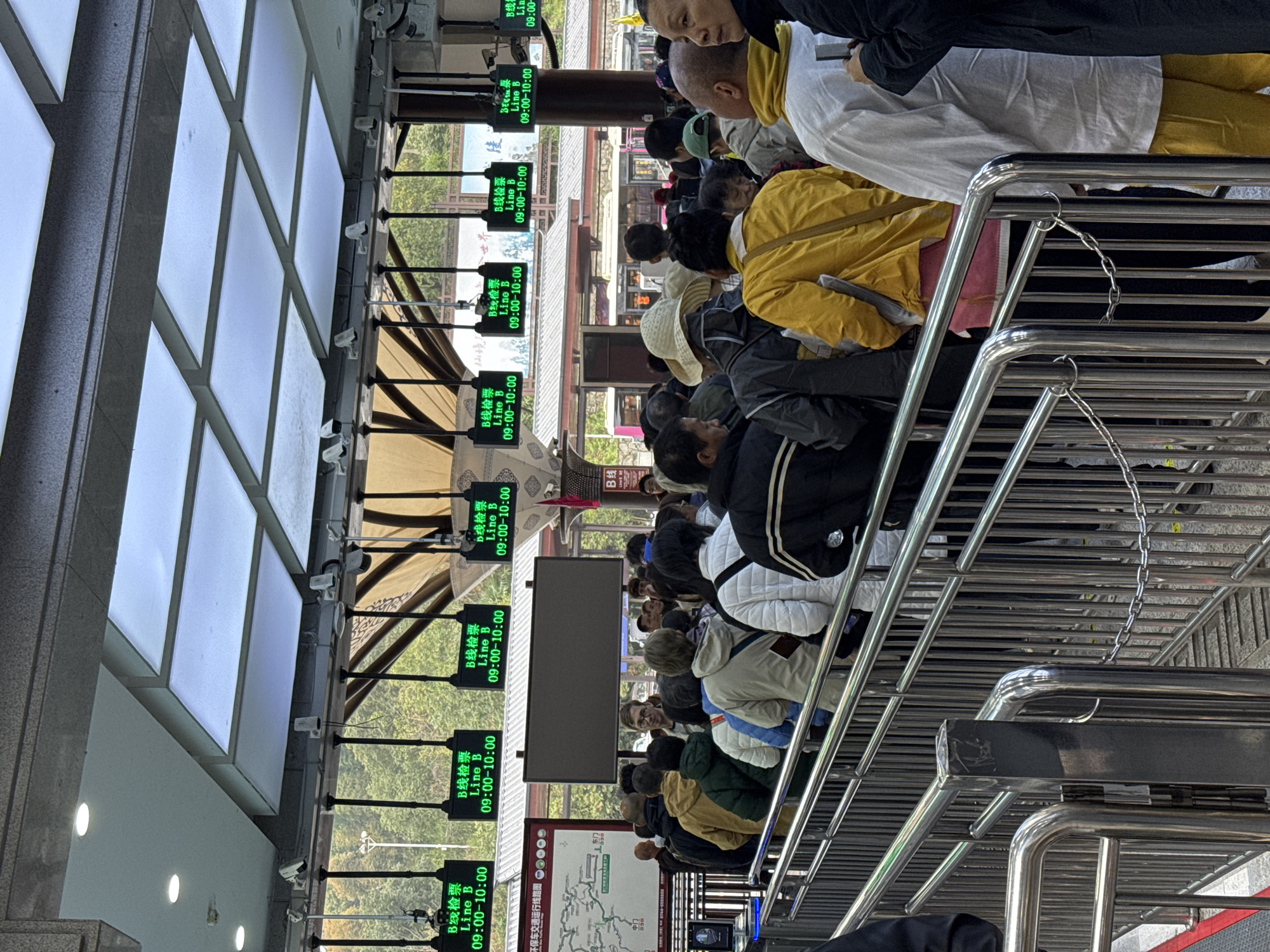 This image captures a crowded outdoor queuing area at a transportation or attraction entry point in Wulingyuan, China. Numerous individuals, estimated at over fifty, are standing in multiple lines formed by chrome-plated metal barriers. Most individuals are viewed from the back or side, dressed in various casual outerwear including jackets and hoodies in colors such as yellow, white, black, grey, and olive green. One man with glasses in the midground is looking directly towards the camera. Another individual on the far right wears a white top, yellow pants, and has a shaved head.
Overhead, multiple green LED screens are suspended, each displaying "B线检票 Line B 09:00-10:00," indicating a timed access line. A large, dark, rectangular display screen, currently blank, is mounted centrally above the crowd. The queuing area is beneath a structural canopy featuring square, backlit ceiling panels and smaller round spotlights. Several white dome-shaped security cameras are mounted on the ceiling structure. To the left, a map or informational sign with Chinese text is partially visible. In the background, beyond the queue, low-rise buildings with red roofs are discernible, with a pink and white bus or tram partially visible further back. Green trees and hills are present in the distant background, indicating a natural setting adjacent to the developed area.