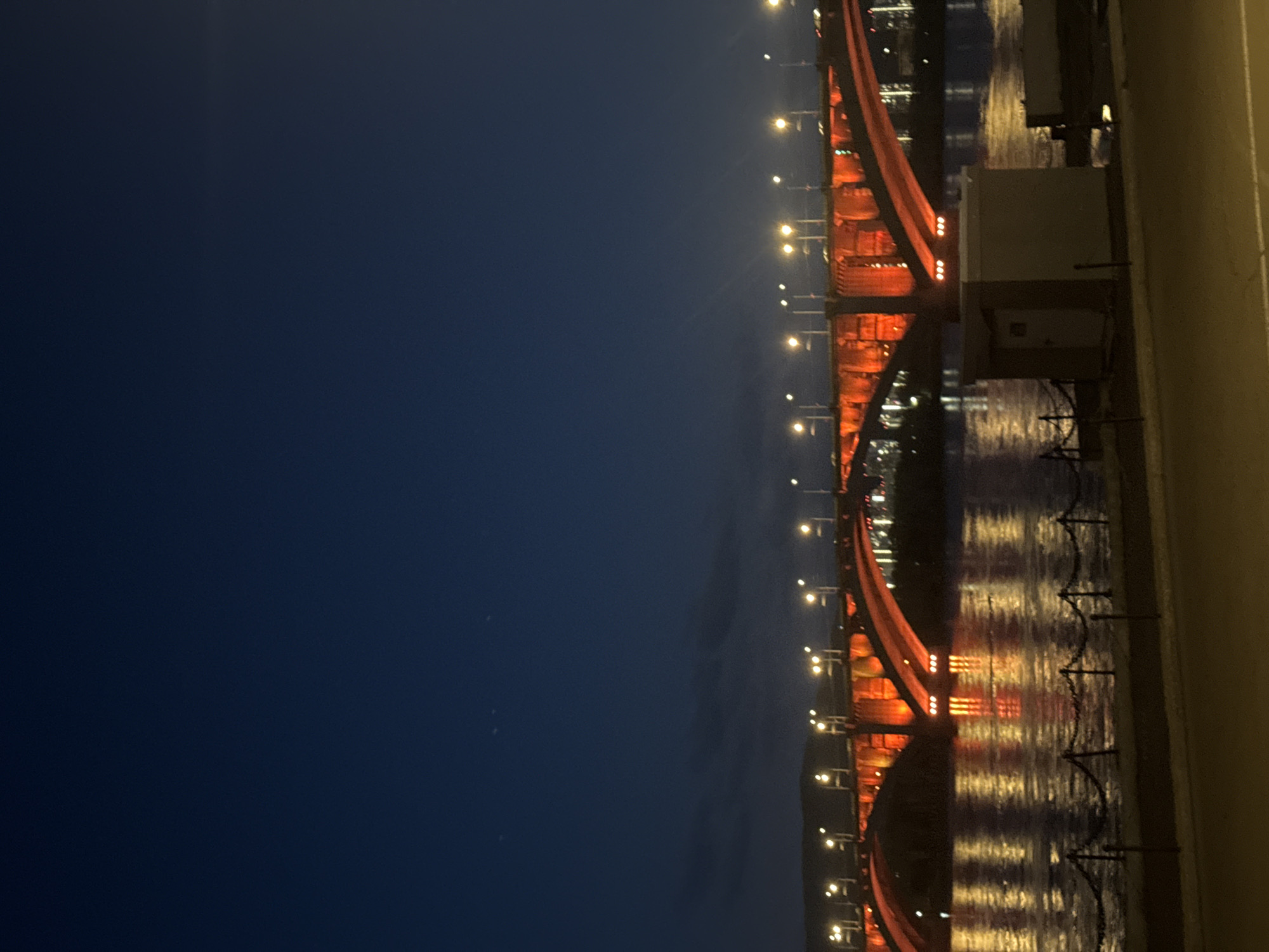 The image shows a bridge over a body of water at night. The bridge is illuminated with orange lights, and the lights are reflected in the water. The sky is dark blue. The image is taken from an elevated position, possibly from another bridge or building. The bridge is located in the city of Krasnoyarsk, Russia.