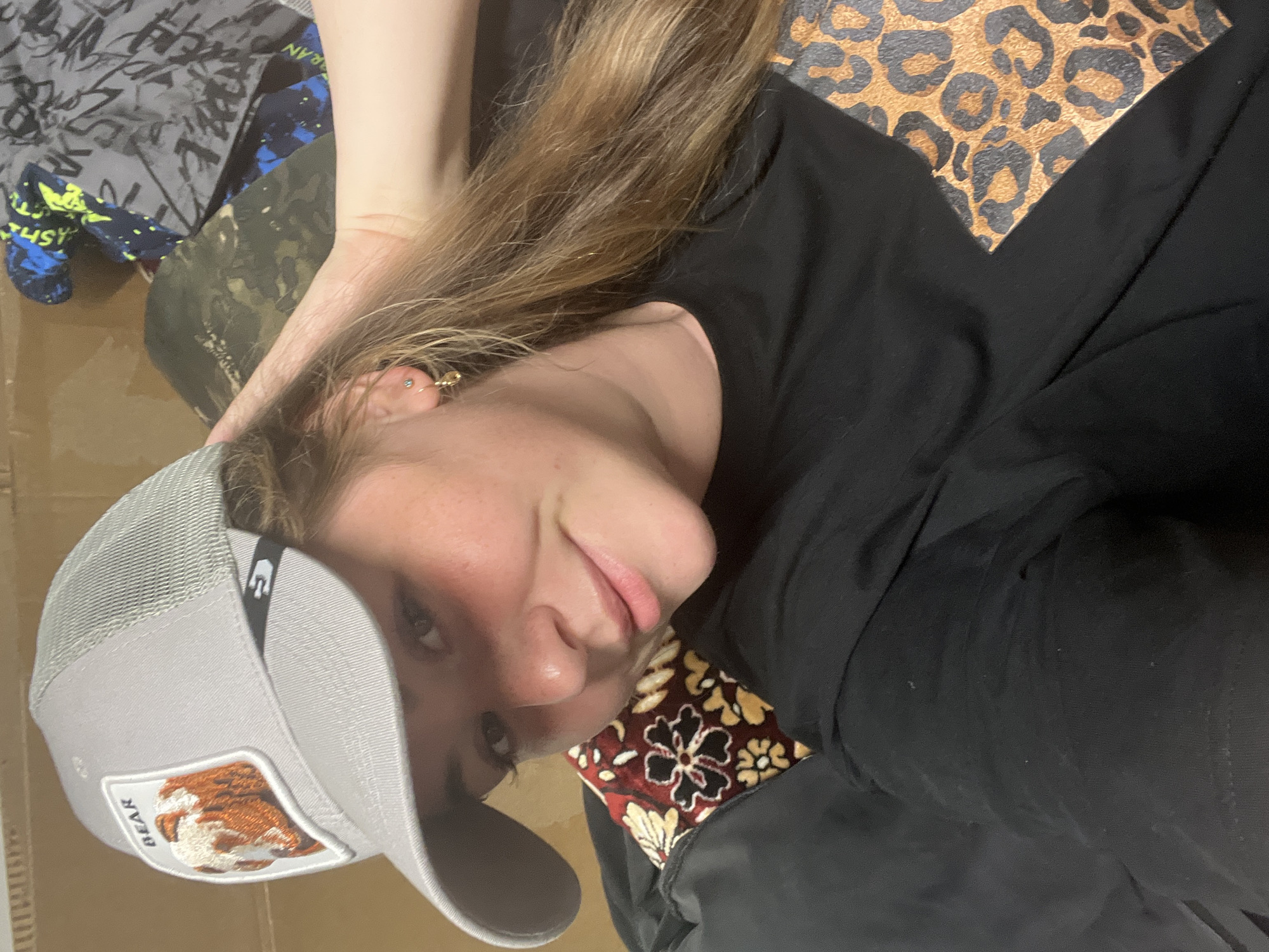 A young woman is lying down and looking at the camera. She is wearing a gray trucker hat with a "Bear" patch on the front, a black long-sleeved shirt with a leopard-print panel on the shoulder and a floral-patterned cuff. Her hair is light brown with blonde highlights. She has multiple piercings in her ear. The background includes various fabrics and what appears to be a cardboard box. There is no location information in the image. The image does not appear to contain any information relating to Chaikovsky, Russia.
