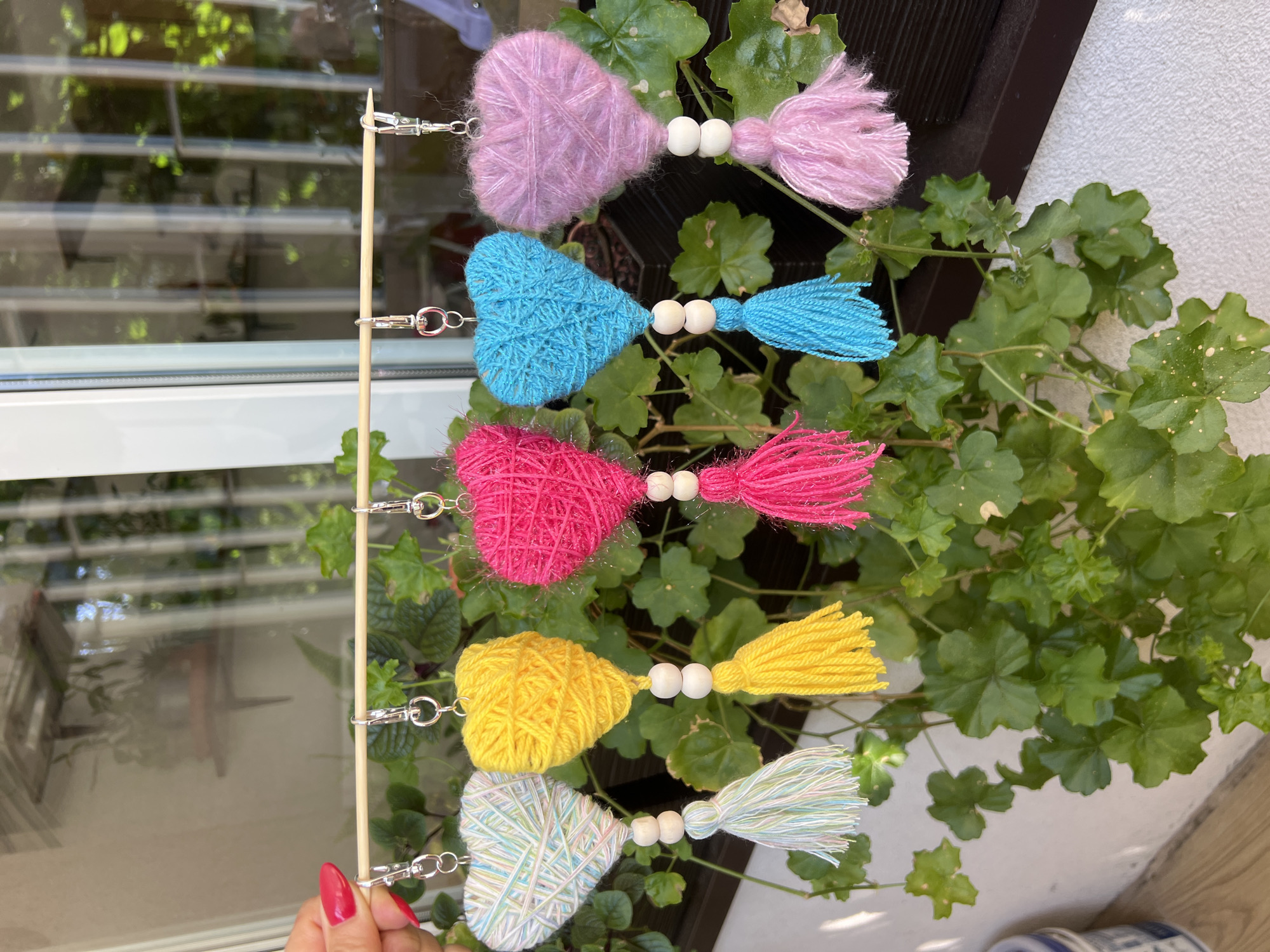 A close-up, eye-level, outdoor shot features five yarn-wrapped heart-shaped ornaments, each hanging from a thin wooden dowel. The ornaments are of varying colors: white with pastel streaks, yellow, hot pink, bright blue, and lavender. Each heart has two wooden beads and a tassel hanging below it. The dowel is held at the left by a hand with bright pink fingernail polish. The background is blurred, showing green foliage and a window with reflections of a building and trees. No specific location in Białystok, Poland is discernible from the image.