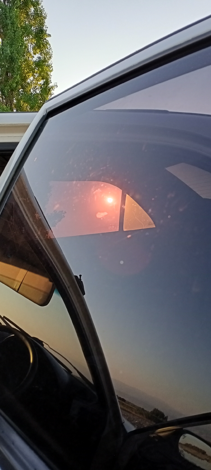 This is a close-up, vertical, eye-level shot of a car's windshield and side window, taken at an unspecified location in Aral, Kyrgyzstan. The image is dominated by the reflections on the glass, obscuring much of the interior and exterior. The most prominent reflection is of the sun, appearing as a bright, reddish-orange orb with some atmospheric haze. This reflection is fractured into two distinct geometric shapes by the car's window frame. The sky, visible through the glass, is a clear, pale blue, transitioning to a warmer, more orange hue near the horizon, suggesting either sunrise or sunset. In the upper left of the frame, a large tree with green foliage is partially visible. The interior of the car is dimly lit, with the steering wheel and part of the dashboard visible in shadow. The side mirror reflects a blurry image of the outside environment. The overall scene is characterized by the interplay of light, reflection, and the geometry of the car's windows.