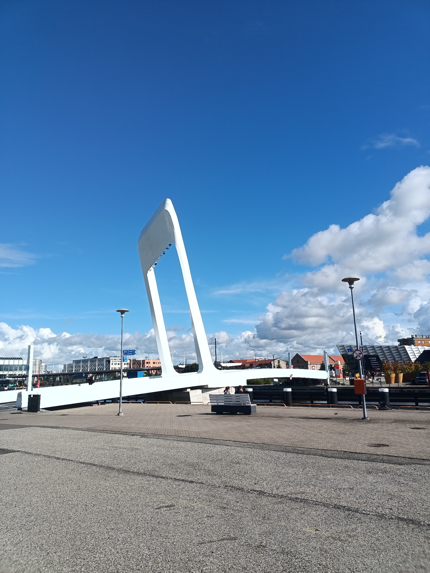 This image depicts a view of the new pedestrian bridge connecting the Old City Harbor to the Linnahall event venue in Tallinn, Estonia. The bridge, a striking white architectural element, is angled upwards, creating a dramatic visual against a bright blue sky filled with scattered white clouds. In the foreground, a paved walkway or road leads towards the bridge. The middle ground shows the bridge itself, with elements of the harbor and surrounding buildings visible. People can be seen on the bridge and near the water's edge. The background consists of buildings typical of a harbor area, including what appears to be a terminal or event space with a distinctive geometric roof design. The overall scene is one of urban infrastructure set against a clear, bright day.
