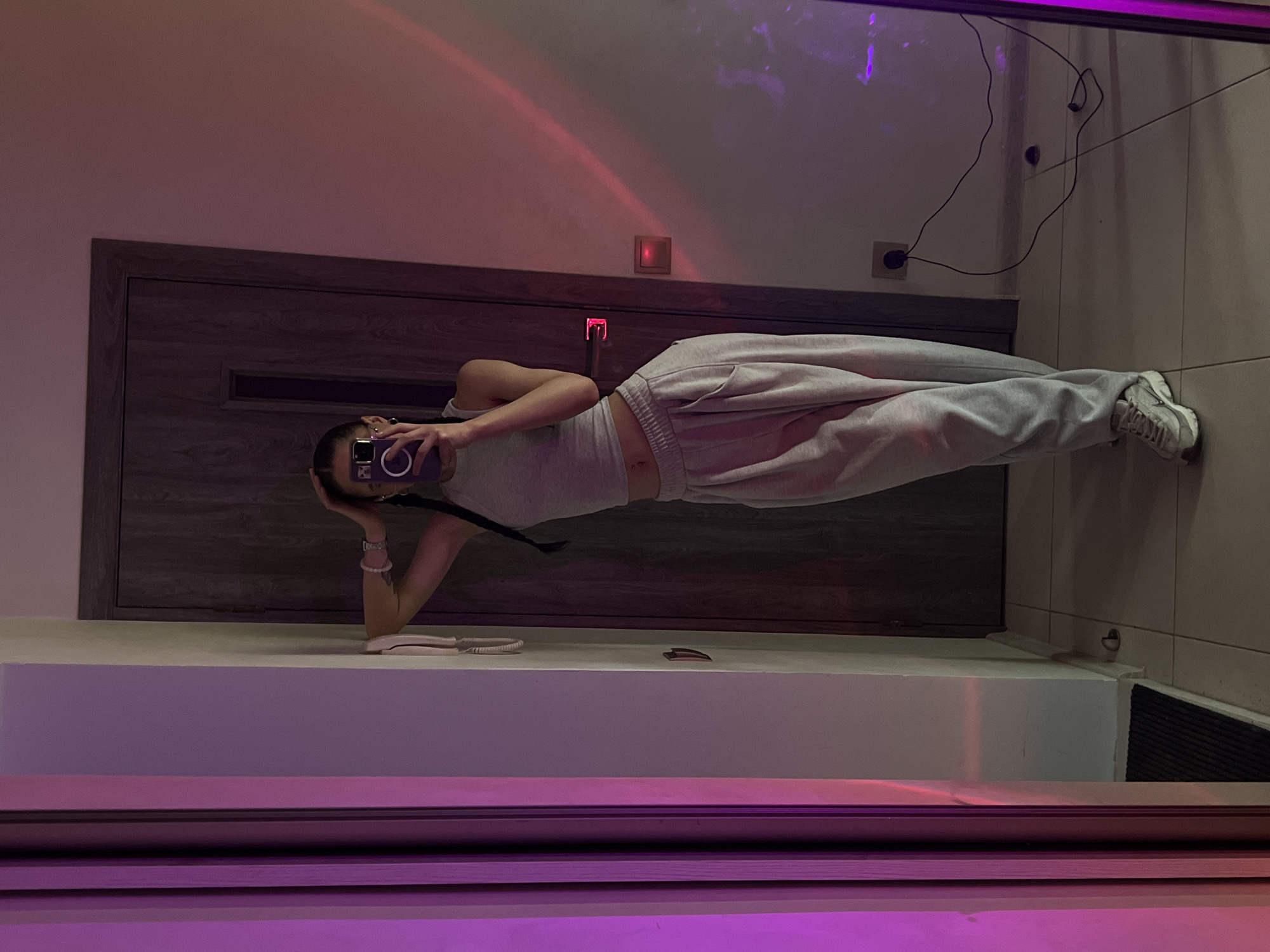The image depicts a young woman standing in front of a full-length mirror, taking a selfie with her phone. She is wearing a grey tank top and light grey sweatpants, with white sneakers. Her hair is styled in braids. The background appears to be a residential hallway or room, with a wooden door on the right and a wall with a light switch and an electrical outlet on the left. The lighting is atmospheric, with streaks of pink and purple light casting shadows on the wall and floor, suggesting the use of colored mood lighting. The floor is tiled. The scene is likely captured indoors. There is no indication of the location being in the city of Ternopil, Ukraine.