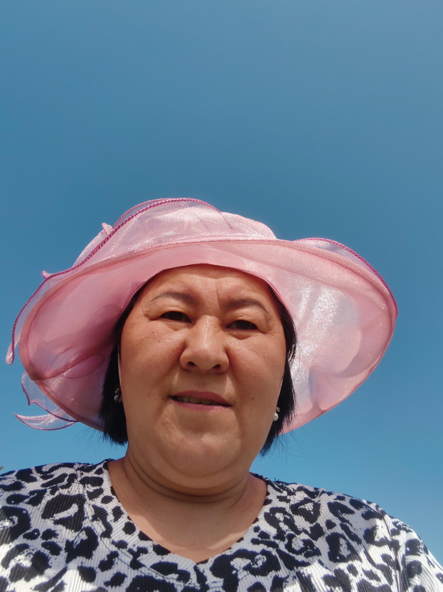 A woman wearing a pink hat and a black and white leopard print shirt looks up at the camera against a clear blue sky. She is outdoors, as indicated by the sky. There are no other people or objects clearly visible in the image. The location of Акжар, Kazakhstan, is not discernible from the visual information provided in the image.