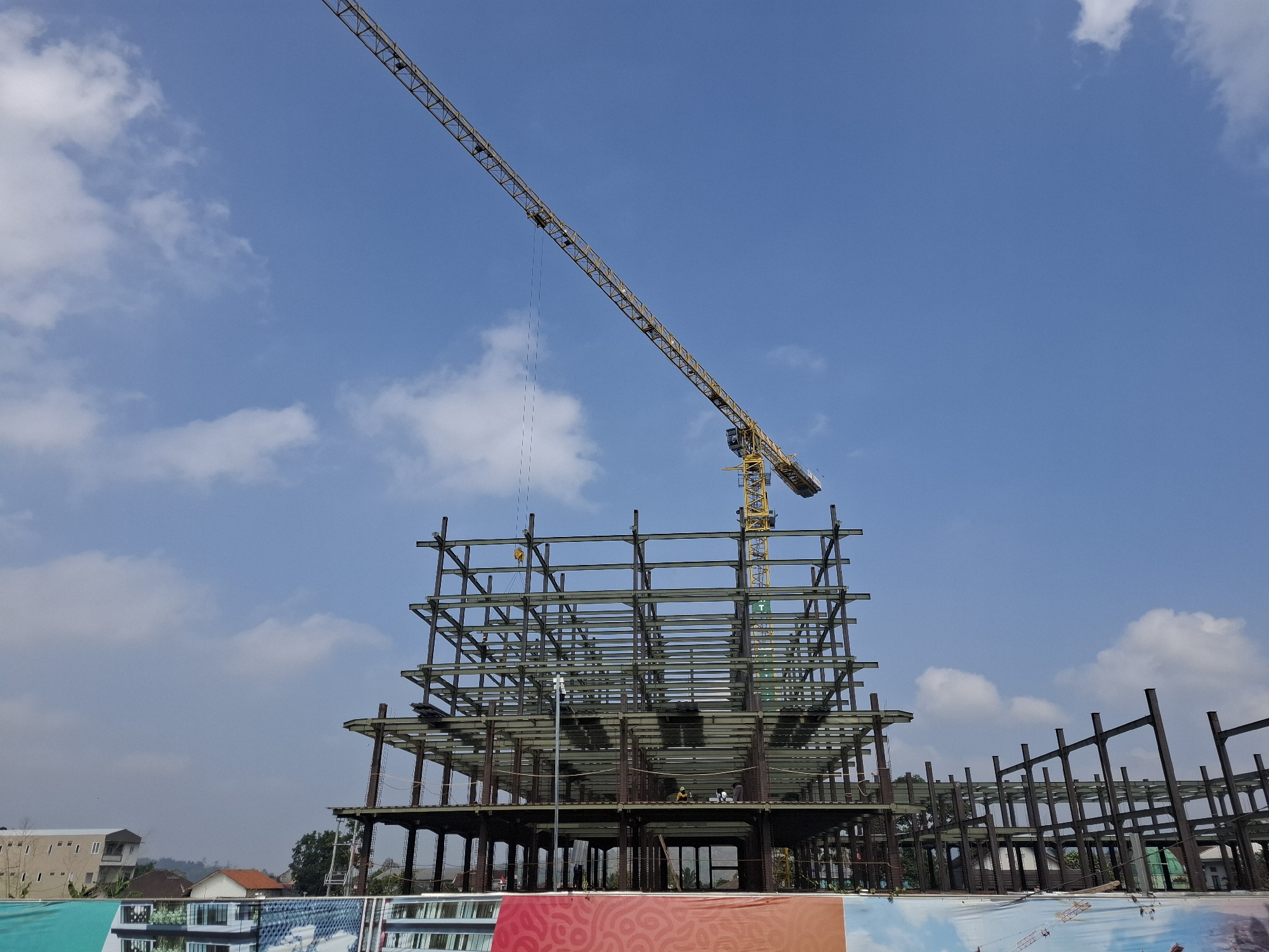 In Selajambe, Indonesia, a multi-story building under construction is visible. A yellow tower crane extends from the right side of the frame, its jib angled upwards into the clear blue sky dotted with white clouds. The building's steel frame is exposed, with multiple levels of horizontal beams and vertical support columns. Several construction workers are present, some appearing to be actively working on the structure at different levels. In the background, other buildings, including a multi-story structure and residential houses with red-tiled roofs, are visible, along with trees. In the foreground, a large banner with abstract colorful patterns is stretched across the bottom of the frame.