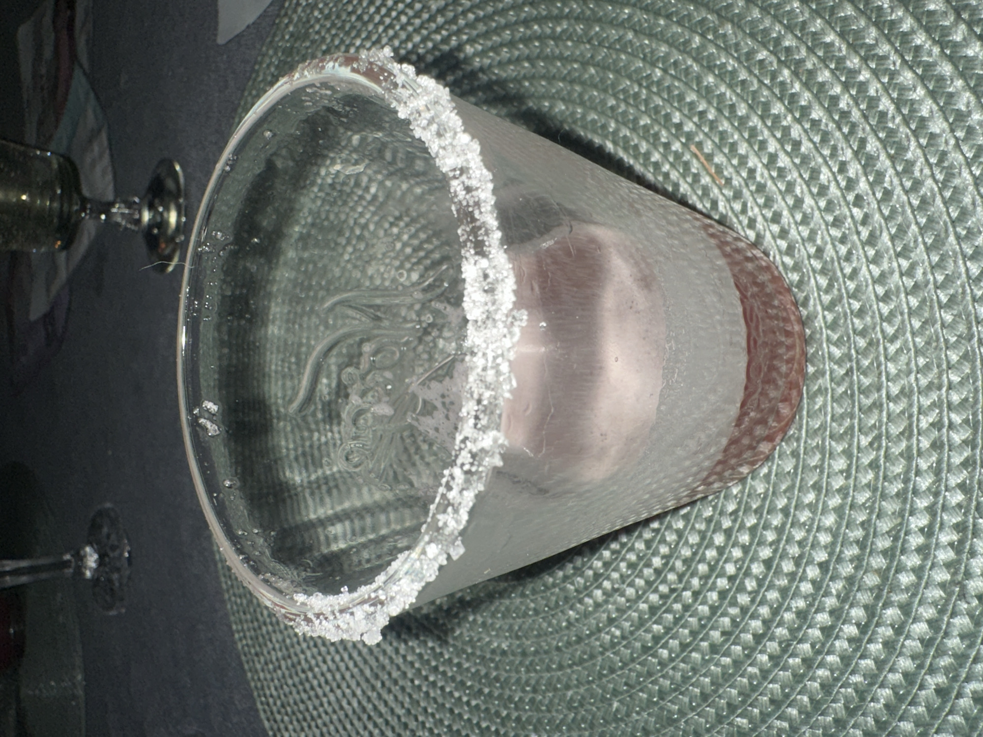A close-up, eye-level shot captures a frosted glass rimmed with coarse sugar crystals, resting on a circular, woven placemat. The glass contains a pinkish liquid, with a visible layer of sediment at the bottom. The background is dark and out of focus, showing a blurred image of a stemmed glass and a dark, textured surface. The image provides no information about the city of Kongsberg, Norway.