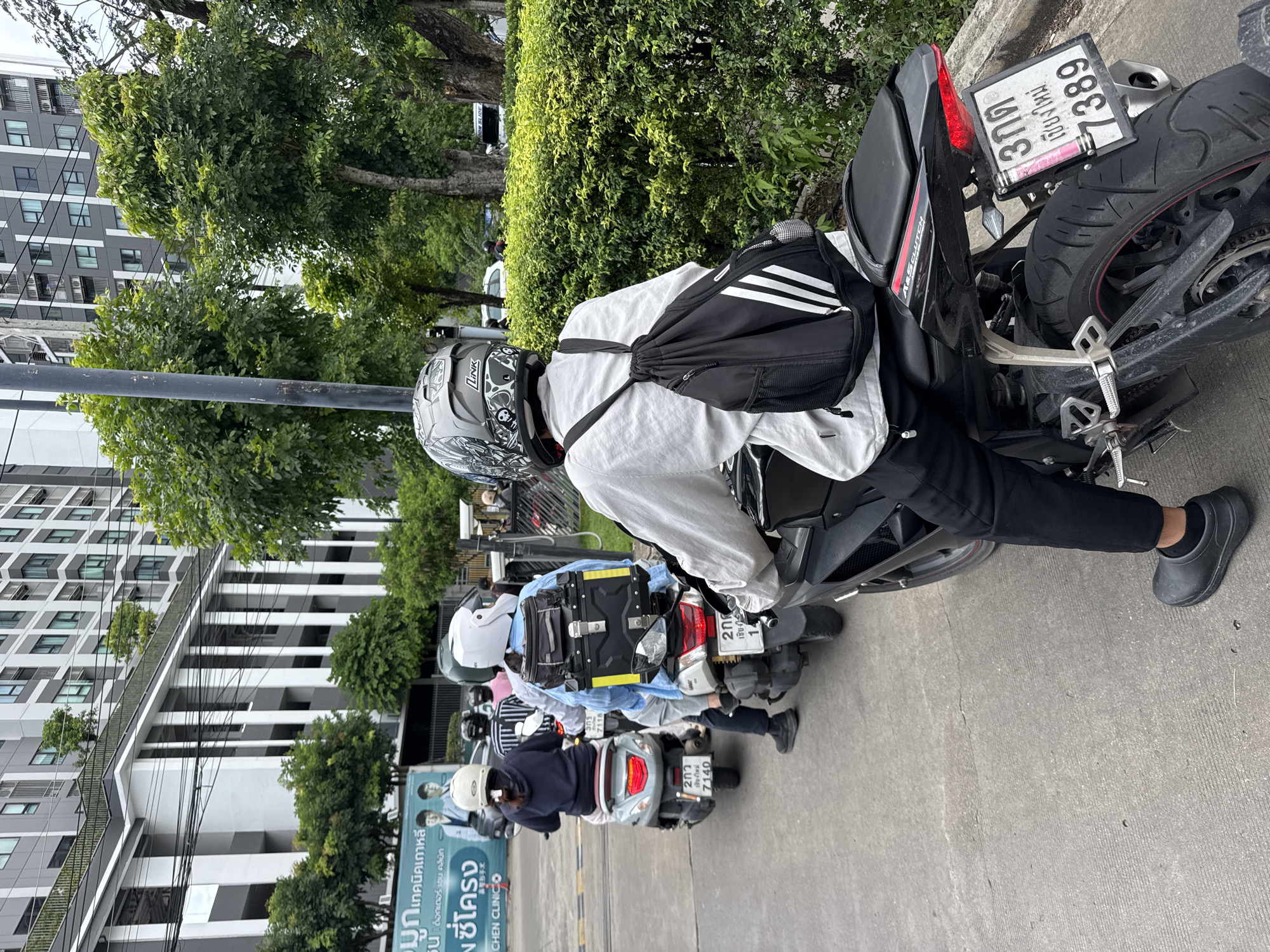 The image shows a line of motorcycles parked on a paved surface, likely a street or parking area, in Thailand. Several people are seen on or around the motorcycles, all wearing helmets. The rider in the foreground, wearing a white shirt, black pants, and black rubber shoes, is leaning against their motorcycle, which has a black backpack slung over their shoulder. Their helmet is grey with a detailed design. The motorcycles are parked in a row, with their license plates visible. One license plate reads "7389" and another "7140". The background features a modern building with multiple stories and many windows, interspersed with lush green trees and bushes. The sky is overcast, suggesting a cloudy day. The overall scene depicts a common mode of transportation in the area, with individuals preparing for or returning from a journey. The text visible on a banner in the background, "มูกเทคนิคเกาหลี ซี่โครง Suด็อกเตอร์ เช่น คลินิก CHEN CLINICO", appears to be an advertisement for a clinic, possibly related to cosmetic procedures or health. The atmosphere seems casual and everyday.