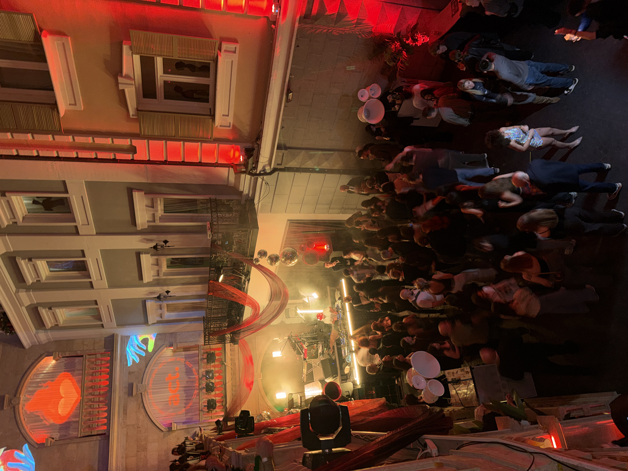 The image captures an outdoor event in Tbilisi, Georgia, possibly a concert or performance, viewed from an elevated perspective. A crowd of people is gathered in a street between buildings. The buildings are illuminated with red light, and one features artistic projections including a heart and a hand. A stage is set up, equipped with lighting and stage equipment. The crowd is a mix of people, some standing and watching the event, others lounging on the ground. There are speakers and decorative elements scattered around.