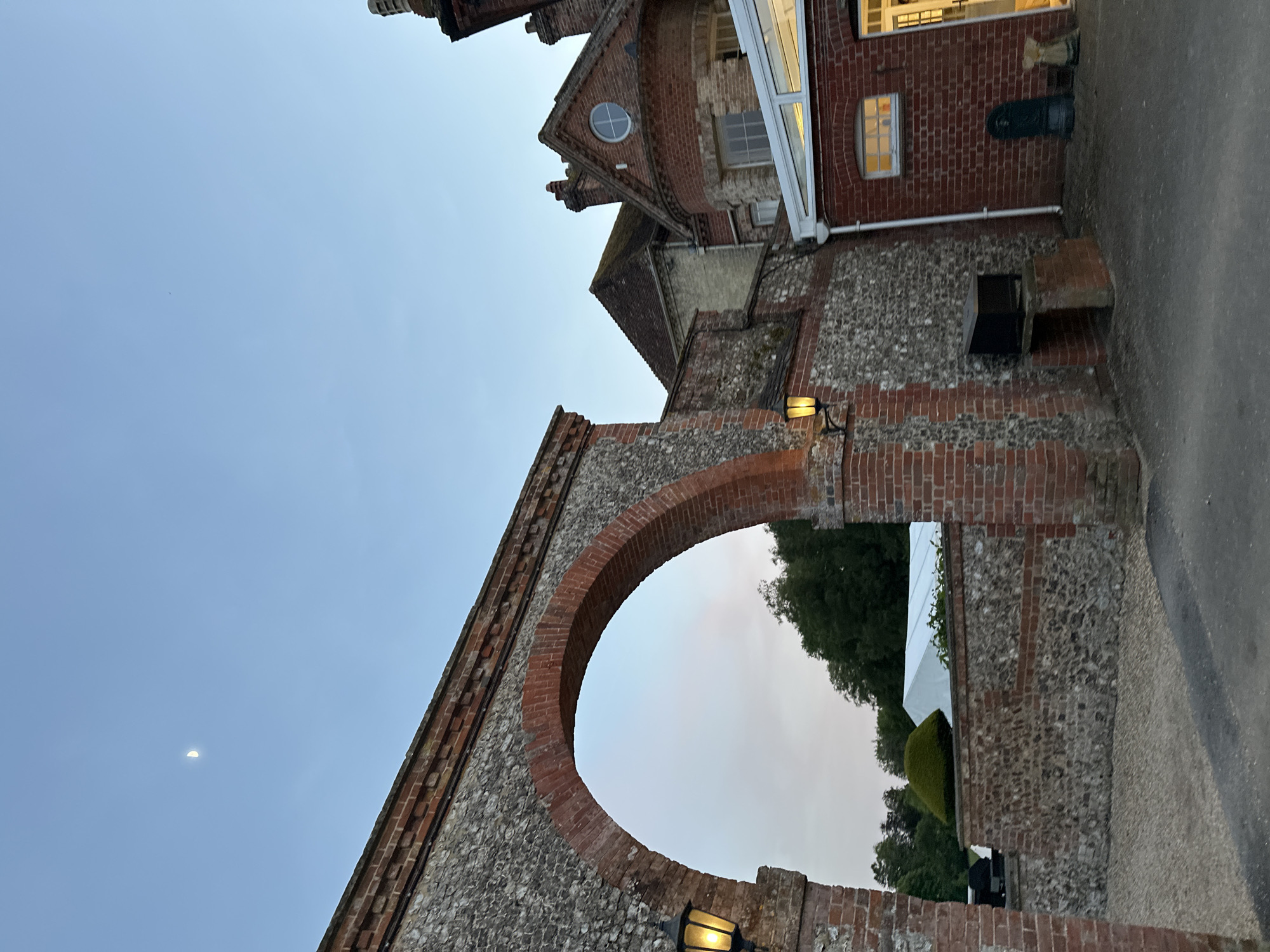A low-angle shot captures an arched stone and brick structure, illuminated by a lamp, framing a view of greenery and a white tent. The sky is a light blue, with a crescent moon visible in the upper left. Adjacent to the archway is a brick building with windows and a small round window. The image is taken from a slightly upward angle, giving a sense of the archway's height. The location is in the city of Unknown, Unknown.