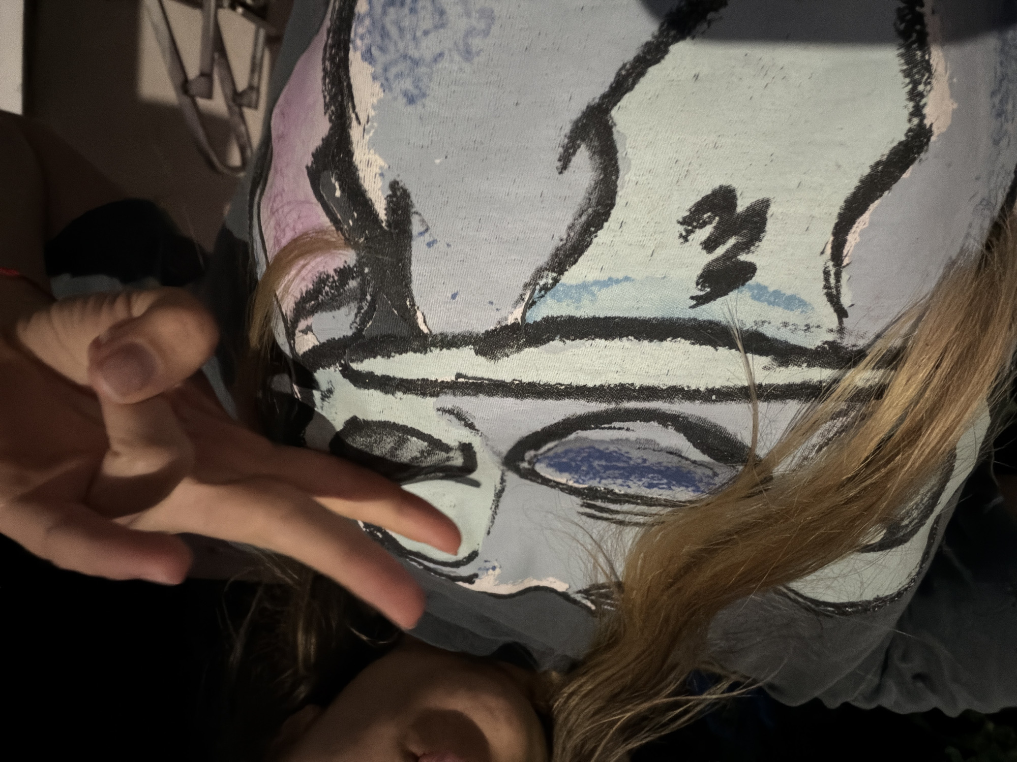 The media file presents a close-up, rotated portrait of a person in an indoor setting in Iskar, Bulgaria, under low-light conditions, possibly evening or night. The image is taken from a slightly upward angle, showing the person's upper body and a hand making a V-sign gesture.
The main subject is wearing a light blue t-shirt adorned with a large, abstract, and stylized face design. The design features bold, dark lines outlining facial contours, eyes (one with blue infill), and a mouth, incorporating shades of black, light blue, and subtle hints of purple. Long, light brown hair cascades over the person's right shoulder and partially covers the t-shirt.
The person's left hand is prominent in the foreground, with the index and middle fingers extended in a V-sign. Only the lower portion of the person's face, including the mouth and chin, is visible at the bottom of the frame, partially obscured by hair and the dark surroundings. The background is dimly lit and indistinct, with some vague outlines of what appear to be metallic or light-colored structural elements in the top left corner, possibly part of furniture.
No visible text can be discerned on the t-shirt or in the background. The overall impression is a casual, posed photograph.