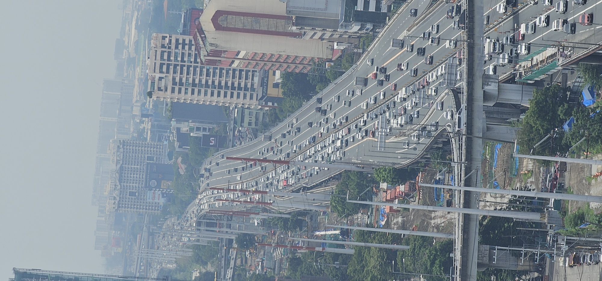 This is an aerial, vertical, panoramic shot of a multi-level highway in Makati, Philippines, featuring a significant traffic jam. The highway is elevated and curves through a densely built urban landscape characterized by tall buildings, some of which are under construction, and various infrastructure elements. The sky is hazy, suggesting air pollution or an overcast day.
On the highway, a dense concentration of vehicles, including cars, vans, trucks, and buses, are visible, moving slowly or stopped. The lanes are filled with traffic. The highway itself is a complex network of elevated roads, ramps, and supports, with multiple levels visible.
Below the highway, on the ground level and in intervening spaces, there are signs of ongoing construction. Construction materials, equipment like excavators, scaffolding, and blue tarpaulins are scattered across the sites. Trees and greenery are interspersed with the urban development, particularly along the sides of the highway and in undeveloped areas.
The buildings in the background vary in architectural style and size, ranging from modern high-rises to older structures. Some buildings have visible signage. The overall impression is one of a busy, developing, and congested urban environment. The focus of the image is primarily on the highway and the traffic, with the city as a backdrop. There are no identifiable individuals or close-up interactions depicted. The scene is dominated by vehicles and infrastructure.
