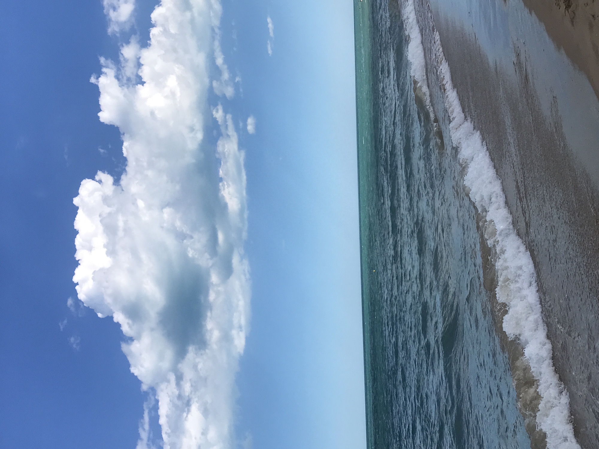 The image depicts a serene beach scene with the Black Sea under a bright, partly cloudy sky. The foreground shows a sandy beach with gentle waves lapping at the shore. The sea itself is a deep blue-green, with white foam cresting on the incoming waves. In the distance, the horizon separates the water from a clear blue sky dotted with large, fluffy white clouds. The scene is devoid of any people or man-made structures, creating a sense of natural tranquility. The location is implied to be Varna, Bulgaria, a coastal city known for its beaches along the Black Sea.