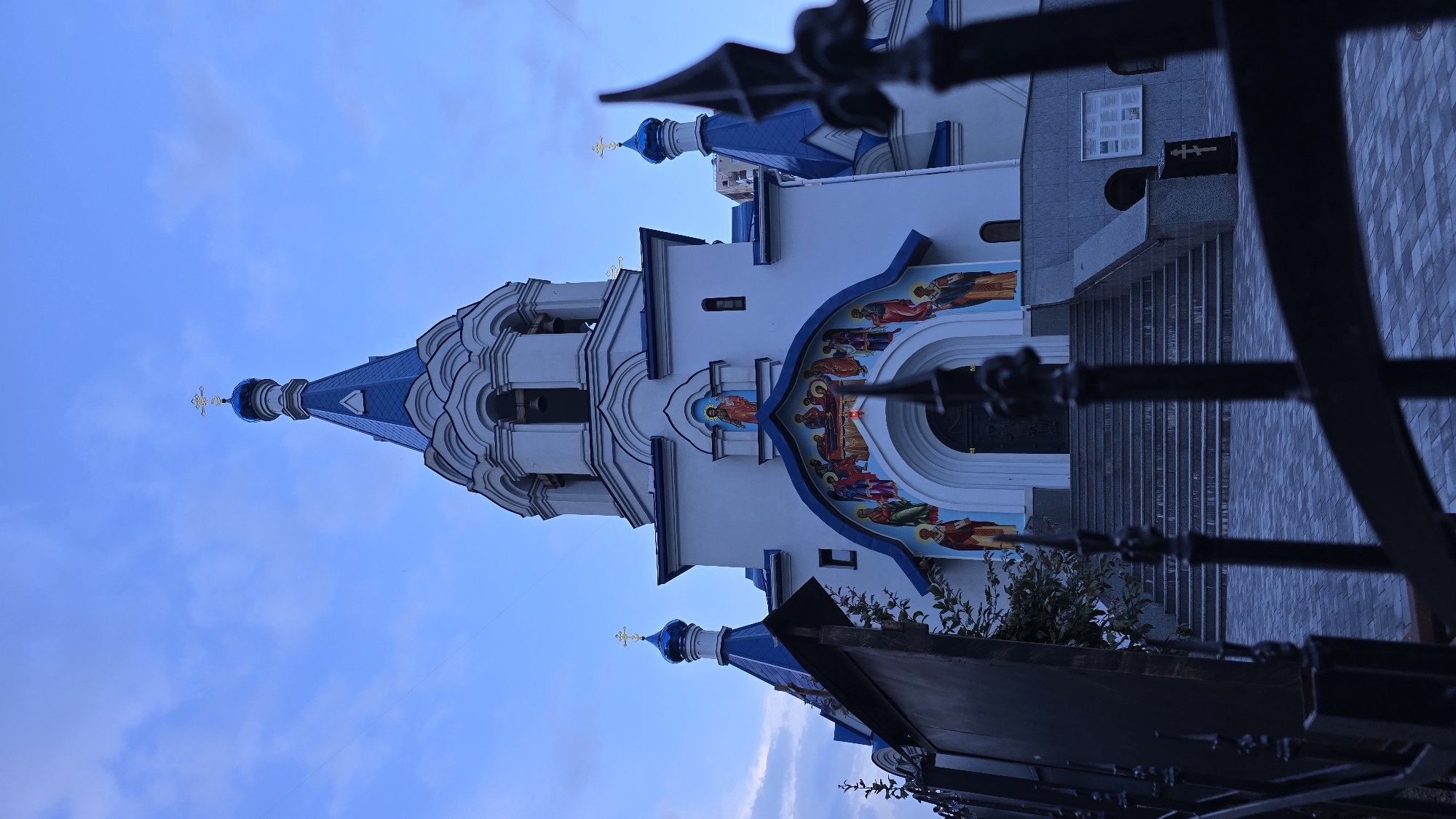 The image depicts the exterior of the Church of the Resurrection of Christ in Kyiv, Ukraine, under a twilight sky. The church features white walls and prominent blue domes, topped with golden crosses. The main entrance is an arched portal adorned with religious iconography, featuring figures of saints or religious figures painted in a vibrant, stylized manner. A set of wide, stone steps leads up to the entrance. A fence with black, ornate metalwork is in the foreground, partially obscuring the view of the church and its grounds. The ground in front of the steps is paved with grey and blue square tiles. The sky is a muted blue with scattered clouds, suggesting the transition from day to night.