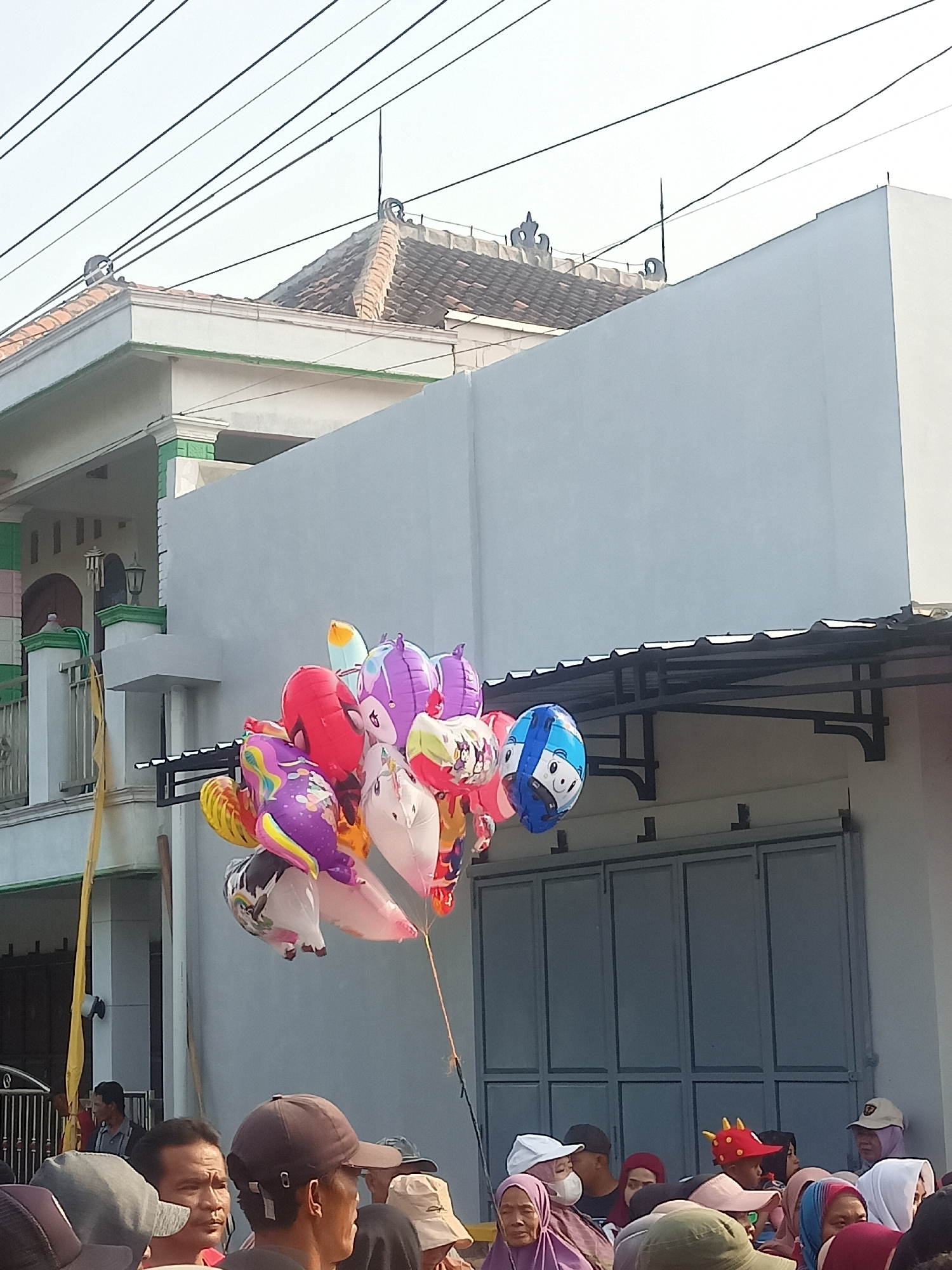In Gemolong, Indonesia, a cluster of brightly colored balloons, some depicting cartoon characters like Spiderman and a blue-faced figure, are held aloft by strings. The balloons are in the foreground, with a light gray building featuring a corrugated metal awning and large grey double doors occupying the middle ground. To the left, a portion of a white and green house with a tiled roof and decorative elements on the roofline is visible. A thin yellow pole stands near the house. A group of people are gathered in the lower portion of the image, some wearing hats and headscarves, suggesting a public gathering or market. The sky is clear and bright, indicating daytime. Electrical wires are strung across the top of the frame.