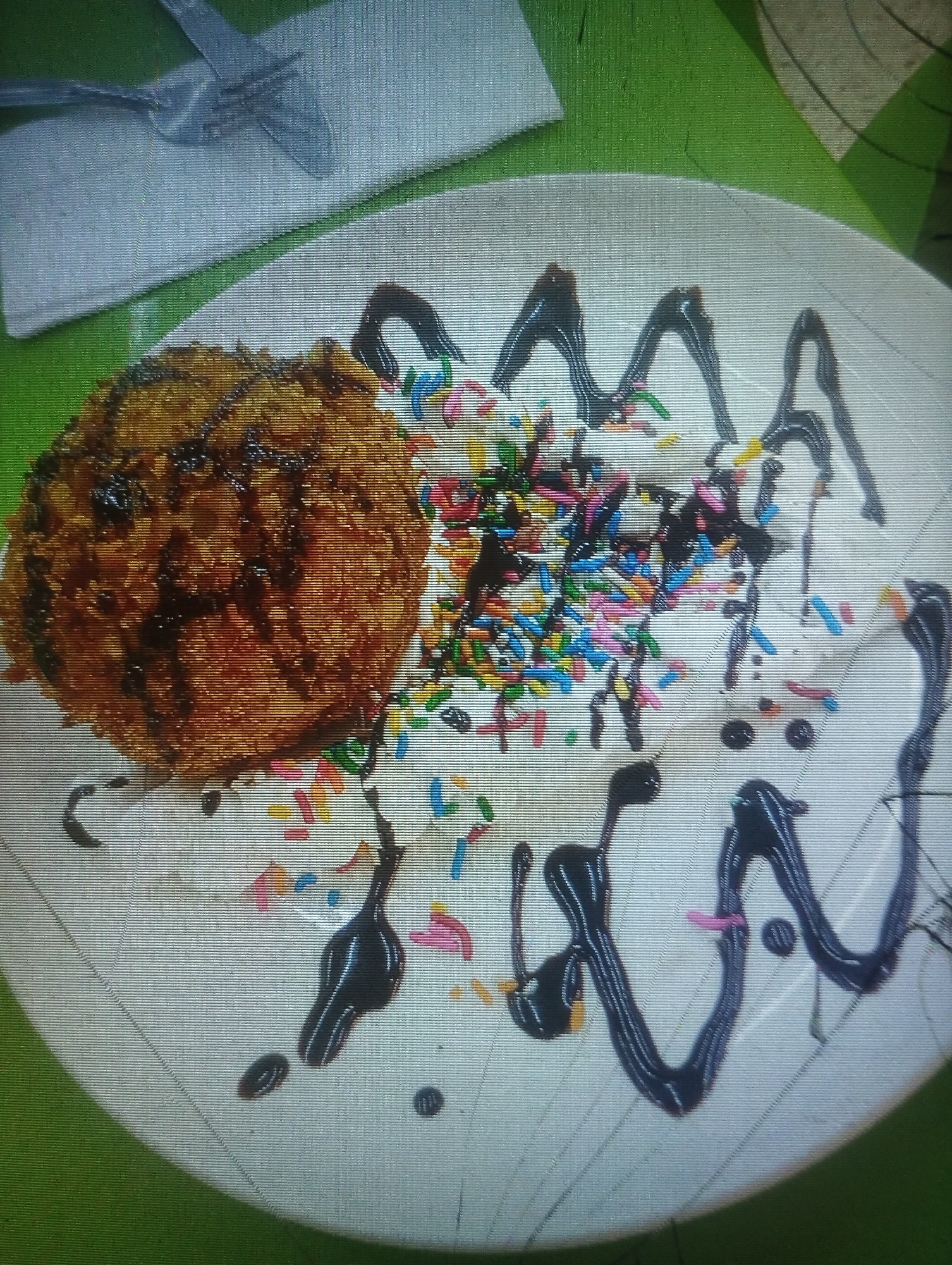 A close-up, slightly angled view captures a dessert dish, likely found in a restaurant in Punta de Piedras, Venezuela. The main feature is a round, fried dessert, drizzled with a dark syrup. It rests on a bed of whipped cream and colorful sprinkles. The dessert is presented on a white plate, decorated with more whipped cream and syrup in a wavy pattern. A fork and knife are visible in the upper left corner, resting on a light-colored napkin. The dessert is on a green surface.