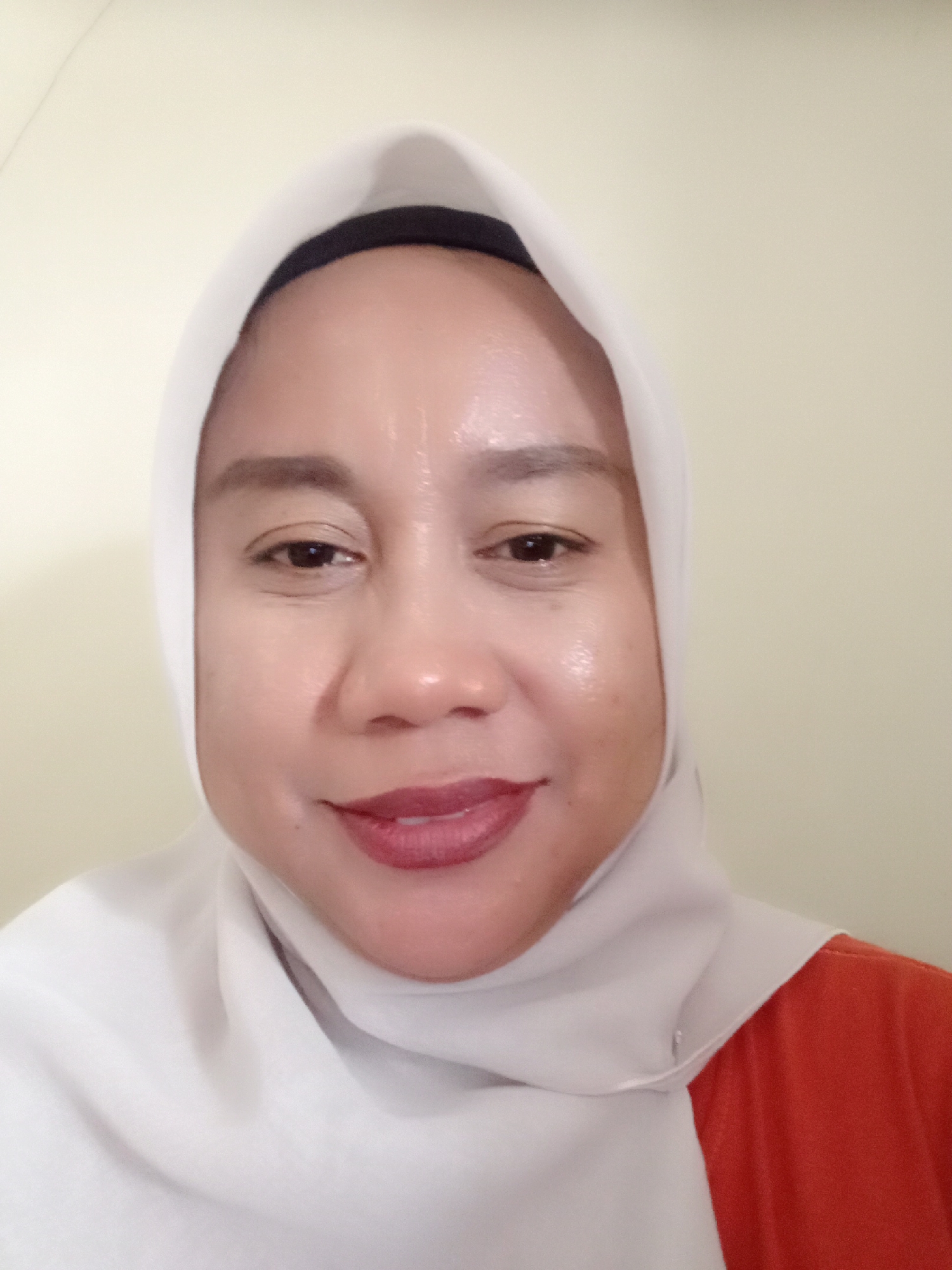A medium shot captures a woman wearing a white hijab and an orange shirt. She is looking directly at the camera with a slight smile. Her face is illuminated, highlighting a dewy complexion and red lipstick. The background is a plain, light-colored wall. The woman's location is stated to be in Wanasalam, Indonesia.