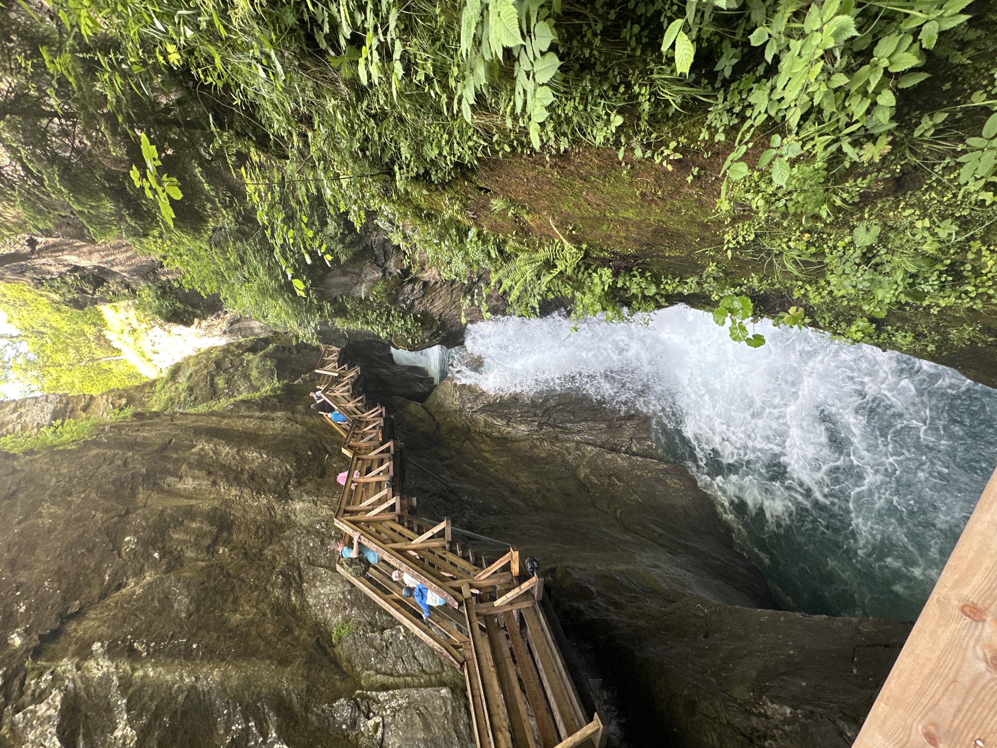 A wooden walkway with railings winds through a narrow gorge in Kaprun, Austria. A powerful waterfall cascades down the right side of the gorge, into a turquoise pool below. Several people, including adults and children, are visible on the walkway, ascending the stairs. The gorge walls are steep and rocky, with lush green vegetation growing from the crevices. The lighting suggests it is daytime, with sunlight filtering down into the gorge.