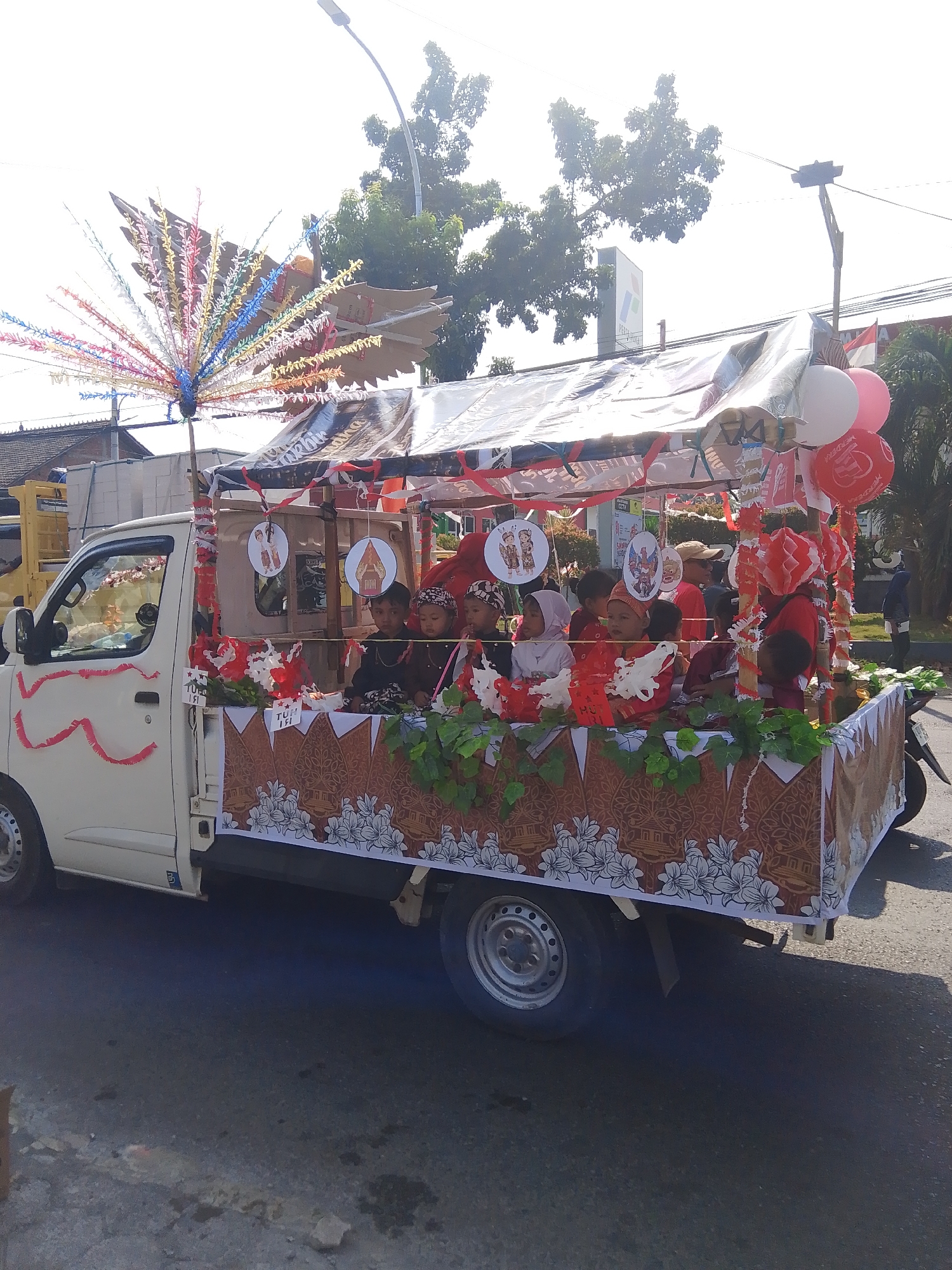 In Rejosari, Indonesia, a decorated flatbed truck is seen in motion, likely participating in a parade or local event. The truck is adorned with red and white streamers and balloons, and its sides feature a patterned fabric with floral motifs and traditional Indonesian house designs. A large, colorful fan-like decoration is mounted on the front of the truck. Several children, dressed in traditional attire, are seated in the back of the truck, which is covered by a makeshift canopy. Some of the children are wearing headscarves. The truck is moving on a paved road, with trees and buildings visible in the background under a bright, sunny sky. A street lamp and a utility pole are also present. The overall scene suggests a festive community gathering.
