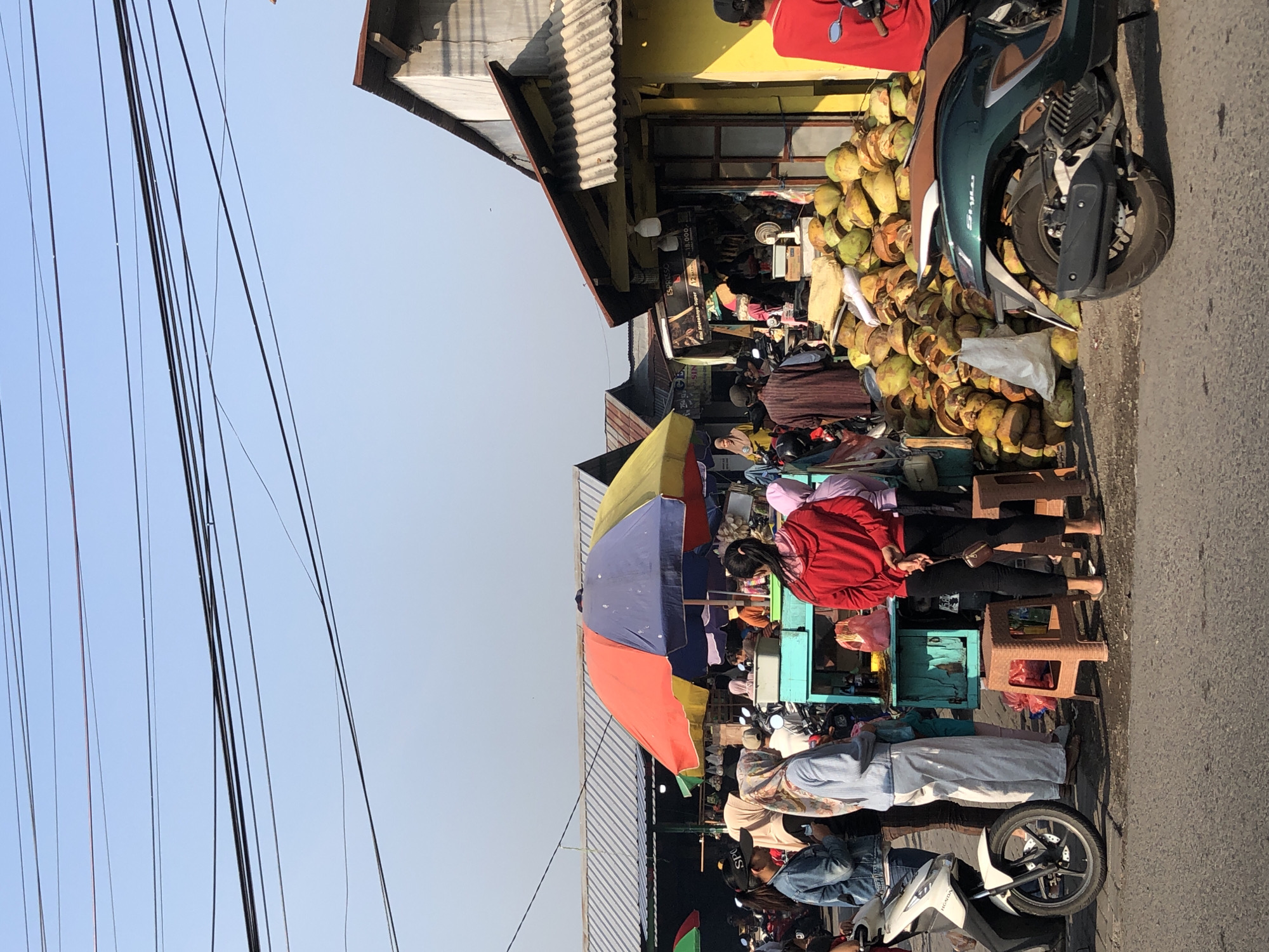 The image depicts an outdoor market scene in Balongdowo, Indonesia, under a clear blue sky with overhead electrical wires. In the foreground, a street market is in progress, with several people browsing and interacting. A pile of coconuts is prominently displayed to the right, suggesting a food or drink vendor. To the left, two motorcycles are parked on the asphalt road.
In the middle ground, a turquoise market stall is set up, covered by a large, colorful umbrella with sections of red, yellow, and blue. Several people are gathered around this stall. Behind it, more market stalls and buildings are visible, some with corrugated metal roofs. A yellow building with a brown trim and a white awning is situated on the right side of the image, adjacent to the coconut display.
The people in the scene are dressed in casual attire. Some are wearing headscarves, indicating a cultural context. One woman in the center is wearing a red hooded jacket and black pants, facing away from the camera. Another woman to her left is wearing a blue denim jacket and a patterned headscarf. A woman further left wears a light blue top and white pants, also with a headscarf. In the background, other individuals are seen, some interacting with vendors or moving through the market. The overall impression is a lively community gathering at a street-side market.