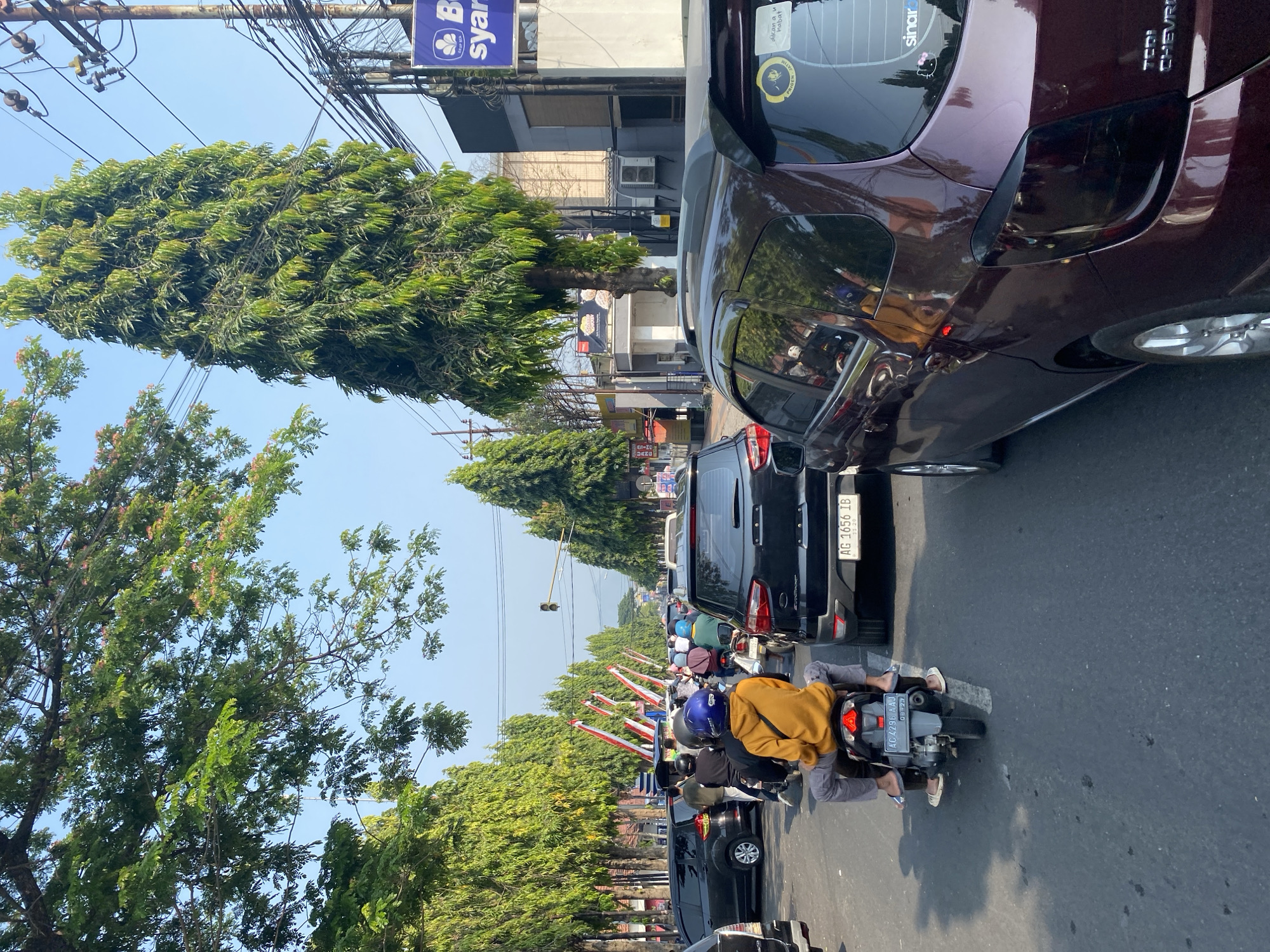 The image depicts a street scene in Kediri City, Indonesia, characterized by moderate traffic congestion. Several vehicles, including cars and motorcycles, are visible on the asphalt road, suggesting a typical urban environment. The presence of red and white flags lining the street indicates a possible celebratory or national event. Tall, slender trees with dense green foliage flank the road, providing shade and contributing to the urban landscape. Overhead, power lines and utility poles are present, common features of city infrastructure. The sky is clear and bright, indicating daytime. A man wearing a yellow hoodie is riding a motorcycle in the foreground, with another person seated behind him. The overall impression is one of a busy street on a clear day in an Indonesian city.