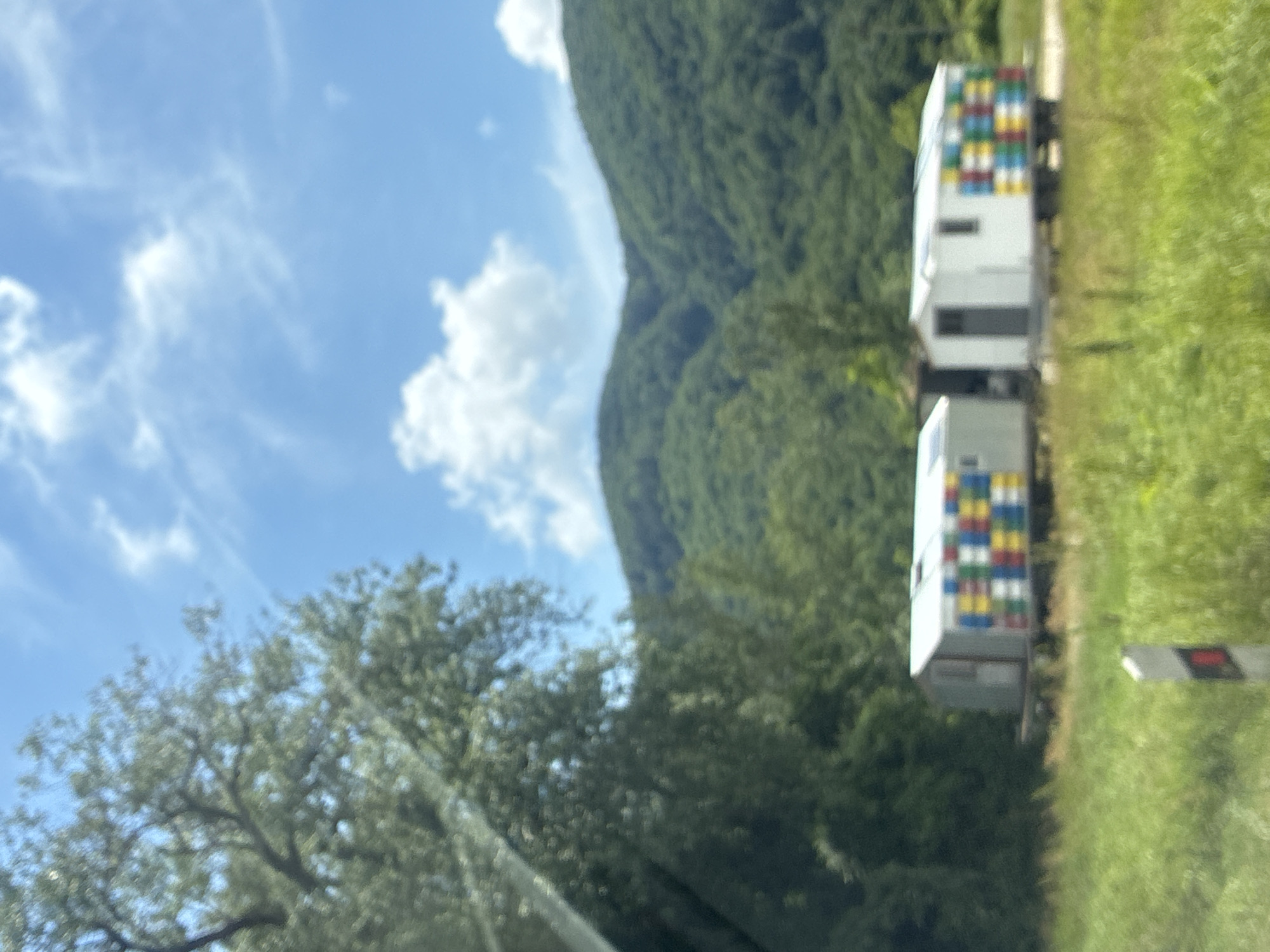 In the city of Tekos, Russia, a scene unfolds under a bright blue sky dotted with fluffy white clouds. Lush green trees and rolling, verdant hills form the backdrop. In the foreground, two mobile structures resembling beehives or small cabins sit on a grassy field. The structure on the left is silver with a gray door and a side decorated with colorful squares of blue, yellow, red, and green. The structure to its right is white with a gray door and a window, and its side also features a grid of the same colorful squares. A simple road or dirt path is visible behind the structures. A white and red road marker stands in the grassy area to the left of the path. The image appears to have been taken from a moving vehicle, as indicated by the slight blur and the presence of what looks like a windshield in the upper left corner.