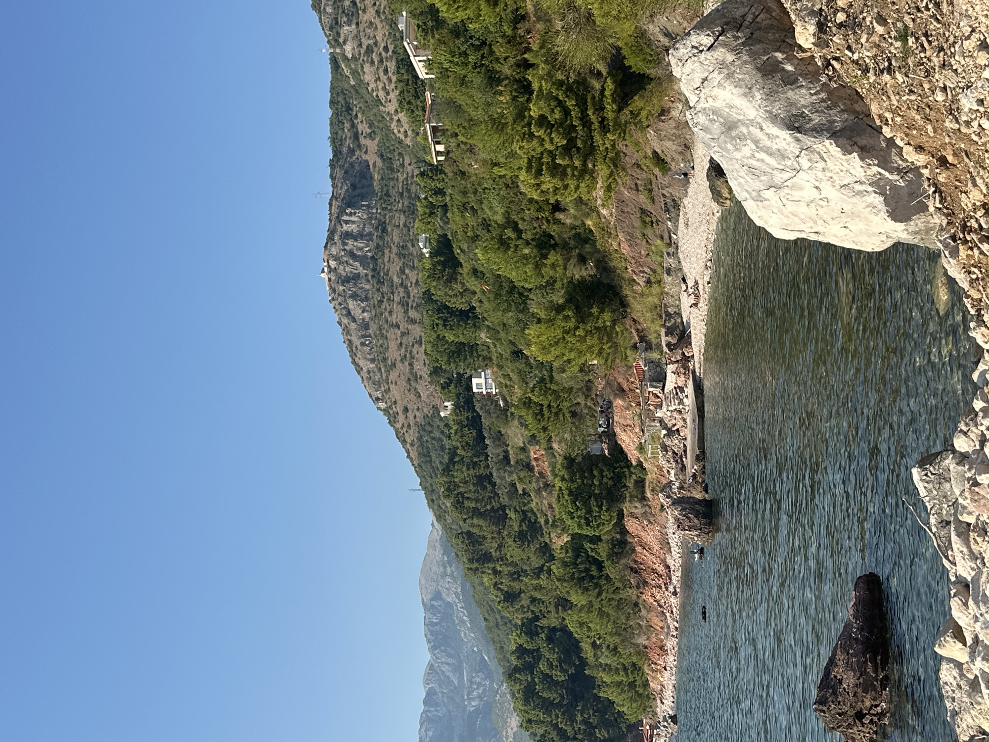 The image shows a coastal scene with a clear blue sky and a calm body of water in the foreground. The water is a deep blue-green and shows ripples on the surface, suggesting a gentle breeze. Several rocks are visible in the water, with some partially submerged and others breaking the surface. The shoreline is rocky and slopes upward, covered with a mix of pebbles and larger stones.
Beyond the shoreline, the landscape is dominated by lush green vegetation, including numerous trees that cover the hillside. The trees are a mix of evergreen species, with dense foliage. Scattered amongst the trees and on the higher slopes are several buildings, appearing to be houses or villas, with white and muted-colored exteriors. These structures suggest a developed, yet natural, environment.
In the distance, a mountain range is visible. The closest mountain to the right is rocky and arid in parts, with some vegetation. The peak of this mountain has a small structure on it, possibly a watchtower or a religious building. Further in the distance, other mountains form a hazy, layered backdrop.
On the beach area, there are some people visible, appearing to be relaxing or sunbathing on the pebbled shore. Some lounge chairs and umbrellas can be spotted, indicating a public beach or resort area. The overall impression is a serene and picturesque Mediterranean landscape. The location is confirmed to be Sveti Stefan, Montenegro.