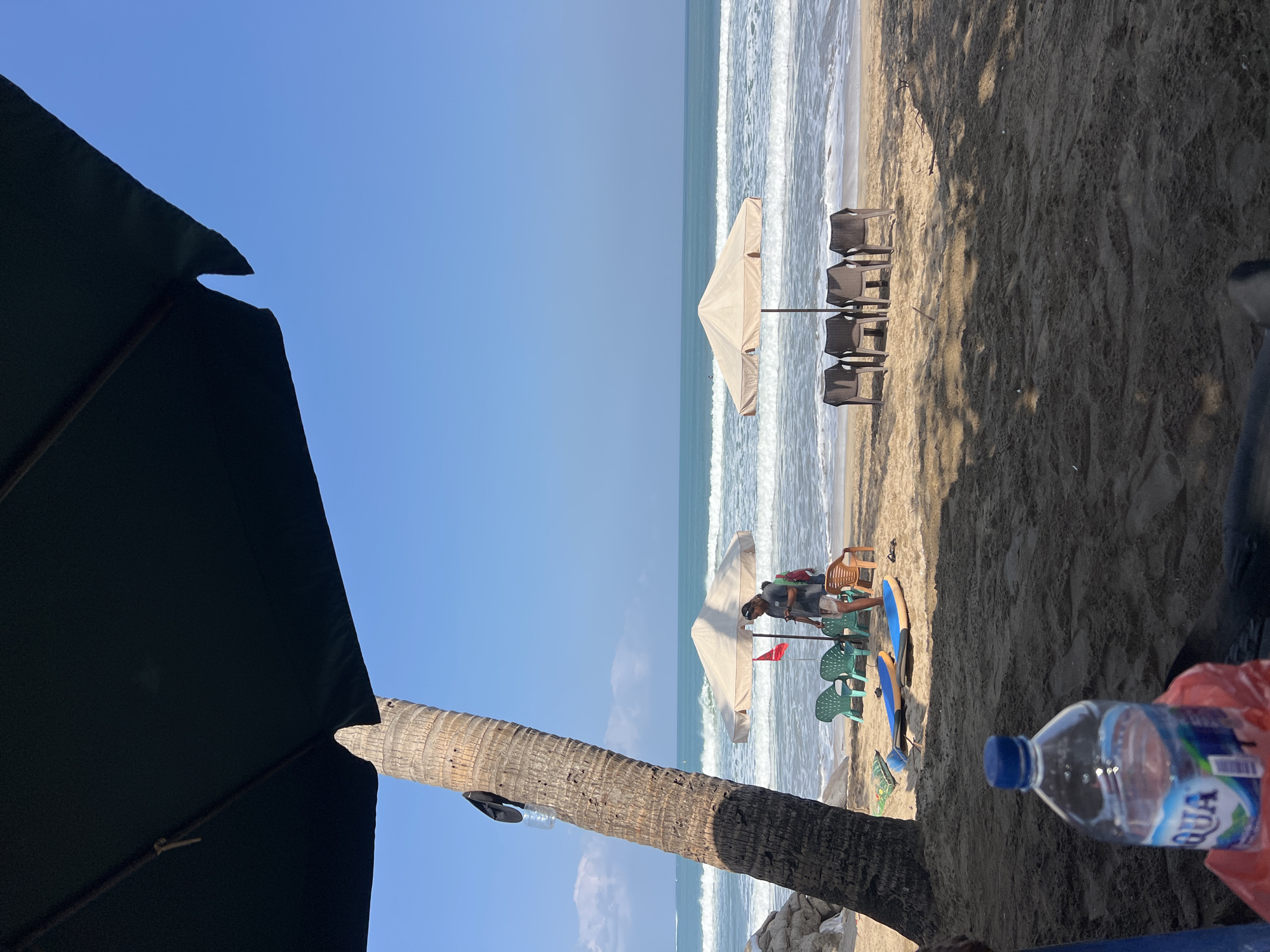 This is a photograph of Kuta Beach in Bali, Indonesia, taken from under a large, dark umbrella. In the foreground, a plastic water bottle is visible on the sand. The beach itself is sandy, with darker sand in the foreground and lighter sand closer to the water. A palm tree with a black hat and a plastic bottle attached to its trunk stands to the left. Further back, several beach chairs are arranged under two separate umbrellas. Two surfboards are lying on the sand near the chairs. A person, appearing to be a man, is standing near one of the umbrellas, looking at a phone. Several waves are breaking on the shore, and the ocean extends to the horizon under a clear blue sky. The overall scene depicts a calm beach environment with typical amenities for beachgoers.