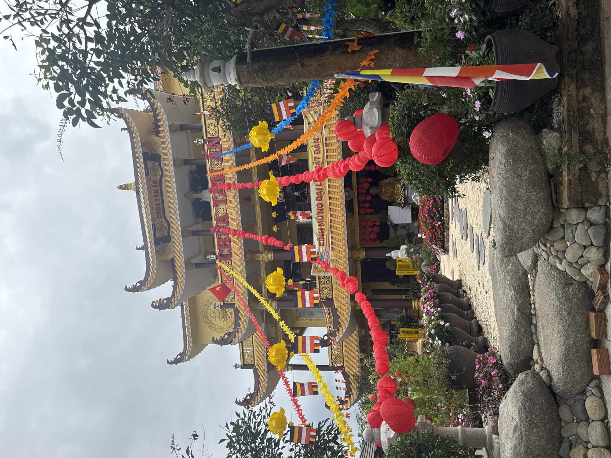 The image shows a Buddhist temple in Da Lat, Vietnam. The temple has a multi-tiered roof with golden accents. The temple is adorned with colorful decorations, including red lanterns, yellow paper flowers, and Buddhist flags. A stone pathway leads to the temple entrance, lined with flowers, greenery, and large stones. The sky is overcast.