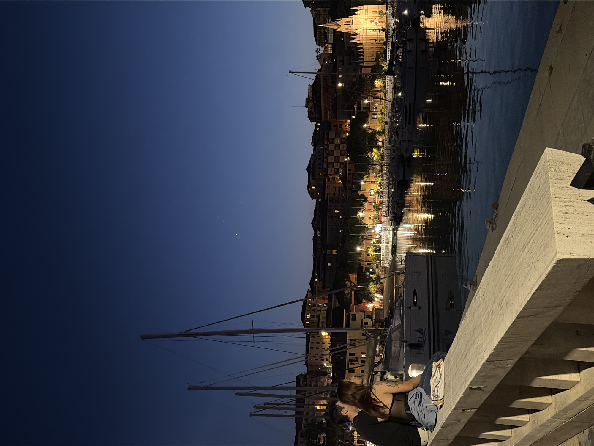 This image captures a nighttime scene in the city of Imperia, Italy, likely at the Imperia Oneglia harbor. In the foreground, a couple is seated on a stone ledge, facing away from the camera and towards the water and the cityscape. The woman has long, dark hair and is wearing a black top and blue jeans. The man has dark hair and is wearing a black shirt. They appear to be in conversation or enjoying the view together.
The background features a harbor filled with docked sailboats and smaller vessels. The masts of the sailboats create a strong vertical element against the skyline. Beyond the harbor, the city of Imperia unfolds, with buildings illuminated by warm lights, creating a picturesque backdrop. A prominent church with a tall spire stands out among the buildings, its facade brightly lit. The dark blue sky above is clear, with a few faint stars visible. The calm water of the harbor reflects the lights of the city and the boats, adding a shimmering quality to the scene. The overall atmosphere is peaceful and romantic, indicative of an evening stroll along the Italian coast.