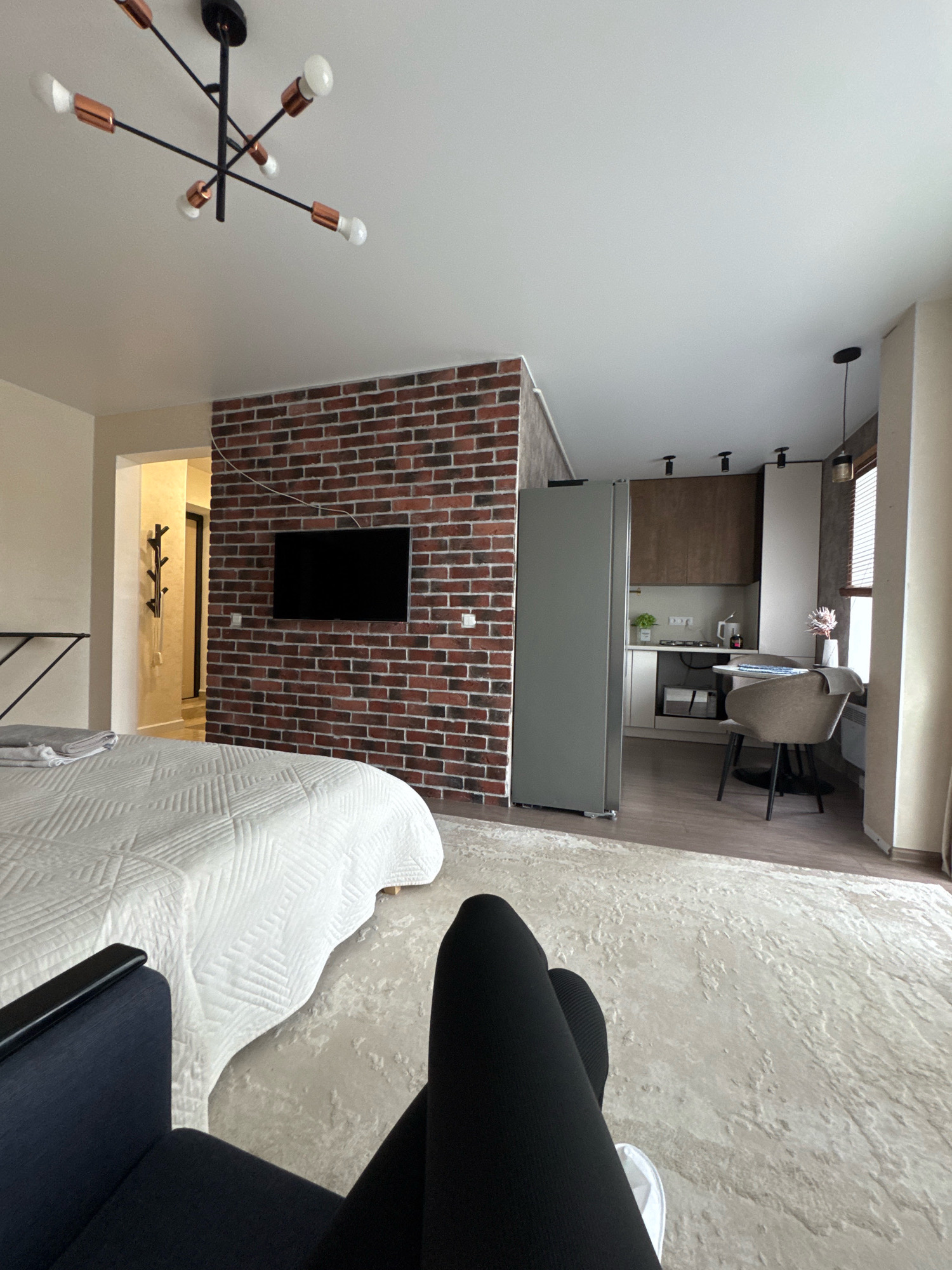 The image depicts a modern studio apartment, likely in Valday, Russia, furnished for comfort and functionality. In the foreground, a person's legs, clad in black leggings and white sneakers, are visible, suggesting they are seated on a dark blue armchair. The armchair faces a large, cream-colored textured rug that covers the floor. Beyond the rug, a white quilted bed is neatly made, with a stack of folded gray towels on top.
The apartment features a distinctive exposed red brick wall, which serves as a backdrop for a black flat-screen television mounted centrally. To the left of the brick wall, an open doorway reveals a hallway with a coat rack. To the right of the brick wall, a compact kitchen area is visible, complete with wooden cabinetry, a white countertop, a stovetop, a kettle, and a small dining table with two chairs. A grey refrigerator stands between the brick wall and the kitchen area. The ceiling is white, and a modern chandelier with copper accents hangs above the bed area. The overall ambiance is one of stylish, contemporary living in a compact urban dwelling.