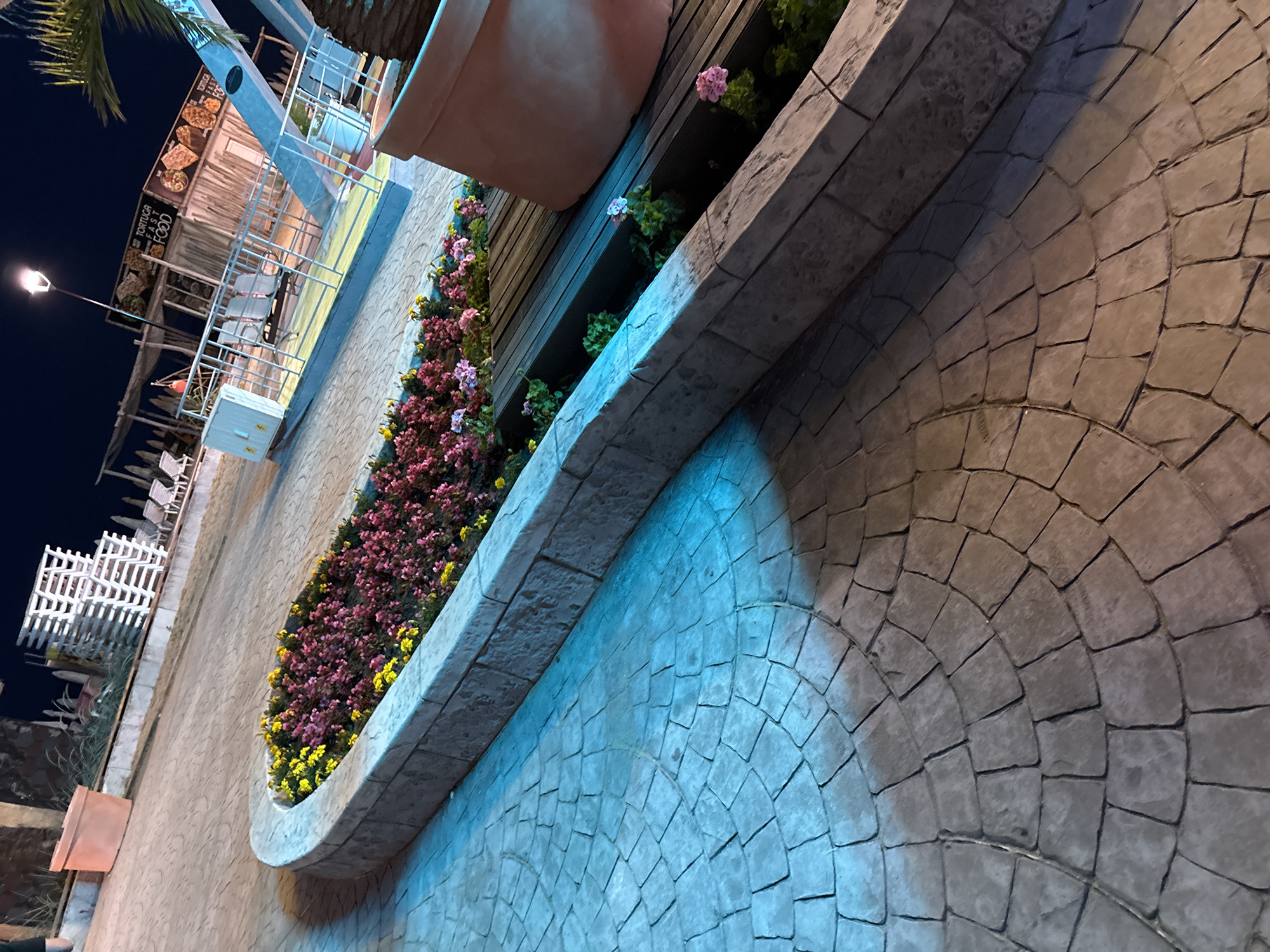 The image depicts a nighttime scene in Varna, Bulgaria, likely at a beachside establishment or entertainment venue. In the foreground, a curved planter bed is filled with an abundance of pink and yellow flowers. The planter itself is constructed from stone blocks. Beyond the planter, a paved walkway with a radial pattern is visible, illuminated by artificial lights casting blue and brown hues. In the mid-ground, a food stall labeled "TORTUGA FAST FOOD" is illuminated, featuring pictures of various food items. Several white chairs are arranged on a raised platform in front of the stall, which is constructed with metallic framing and wooden siding. In the background, stacks of white lounge chairs are visible, suggesting a beach or pool area. A palm tree is partially visible on the right side of the image. The overall atmosphere suggests a recreational or dining area open during the evening.
