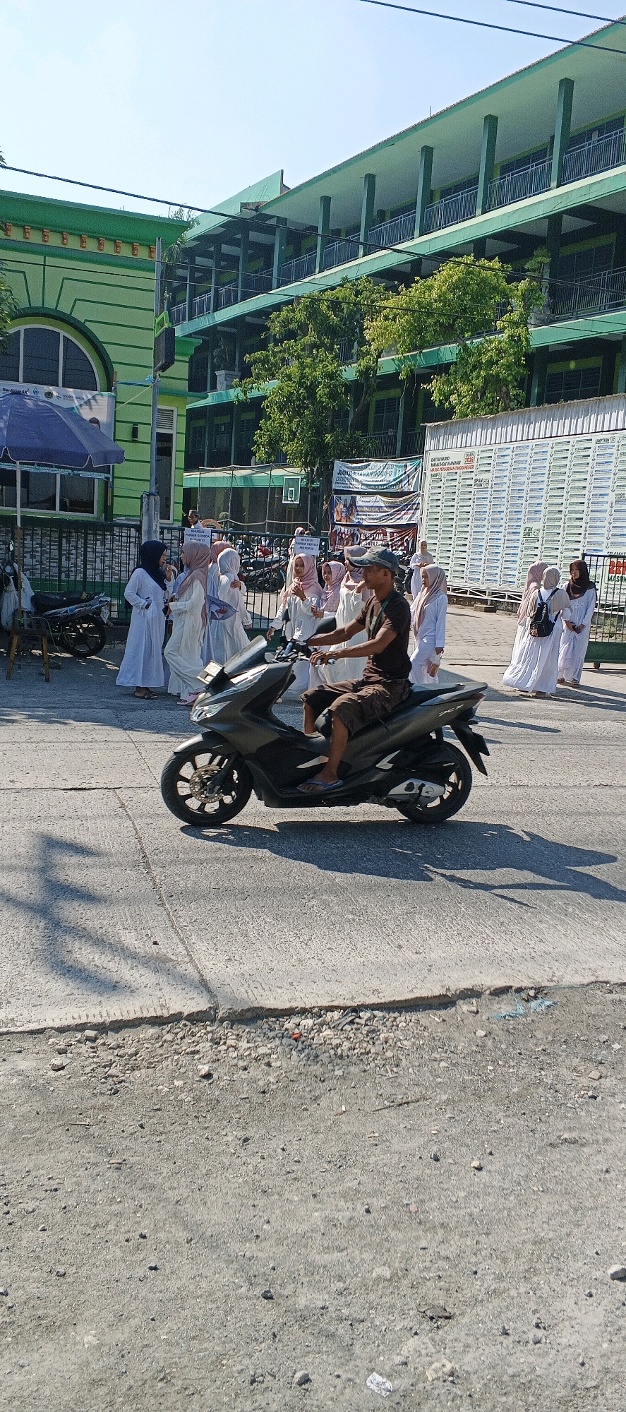 In Sungelebak, Indonesia, a man rides a dark gray scooter down a paved road on a sunny day. He is wearing a brown t-shirt, brown shorts, and a dark baseball cap. Behind him, a group of people, mostly young women in white hijabs and dresses, walk on the sidewalk. A large, green building with multiple levels and balconies dominates the background. Trees and shadows are visible. The road surface is a mixture of asphalt and rougher, gravelly material. Several signs are posted near the building.