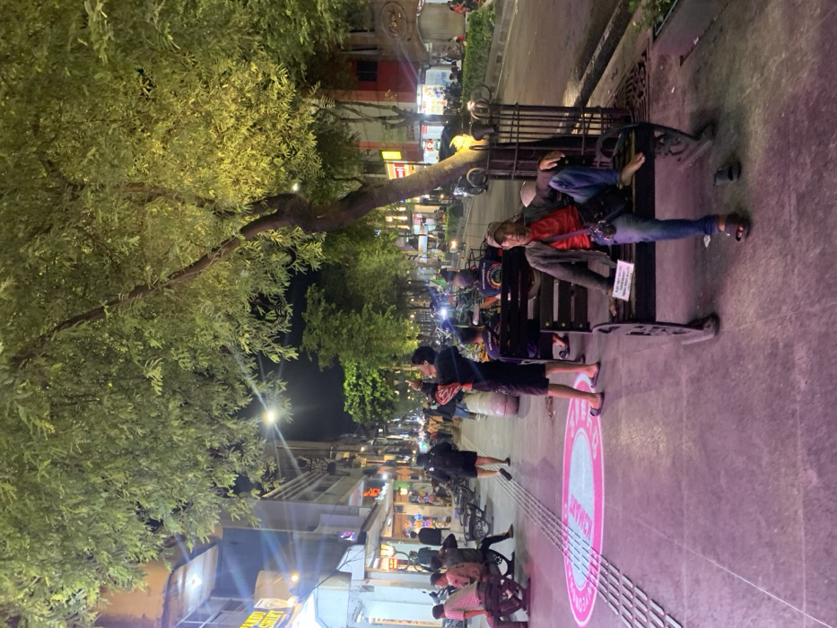 This image depicts Malioboro Street in Yogyakarta, Indonesia, at night. The scene is illuminated by streetlights and shop lights, casting a mix of bright and shadowed areas.
In the foreground, a wide, paved pedestrian sidewalk extends from left to right, featuring tactile paving strips. A circular pink graphic is visible on the sidewalk, with text partially readable as "SELAMAT DATANG" (Welcome).
On the right side of the foreground, a man sits on an ornate dark metal bench. He wears a dark jacket over a red t-shirt, blue jeans, and a light-colored baseball cap. His left foot is in a dark sandal, while his right foot is bare and rests on the bench. A dark shoulder bag is visible across his body, and he holds a small white rectangular sign. Directly behind him, a tree with a gnarled trunk and lush green foliage stands, illuminated by an upward light source.
Towards the mid-ground, slightly to the left of the center, another man stands facing right, looking upwards, with his arms raised as if holding a device to photograph or film something above him. He wears a dark t-shirt and dark shorts, with light-colored flip-flops. Further left, several other individuals are present, some sitting on benches or parts of what appear to be traditional becak (cycle-rickshaws).
The background features a row of multi-story commercial buildings lining the street, displaying illuminated signs such as "PANDU SHIRT." A multi-lane road runs parallel to the sidewalk, populated by vehicles, including cars and motorcycles, and additional pedestrians. More illuminated trees are visible further down the street. Overhead, streetlights emit bright light into the dark sky.