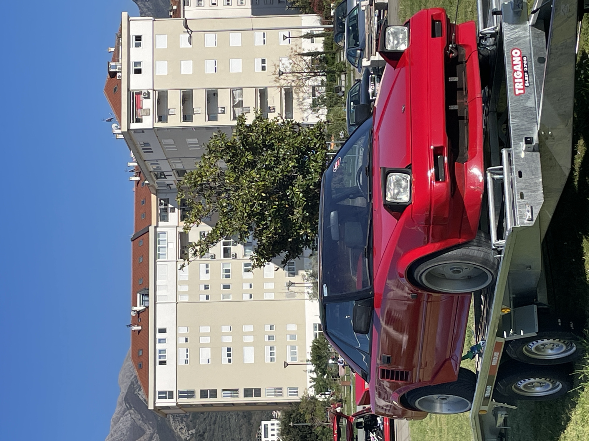 A bright red Toyota MR2 is parked on a silver Trigano trailer, viewed from a slightly oblique frontal angle. The car faces towards the viewer's right. The trailer's hitch is visible. In the background are multi-story residential buildings in Bar, Montenegro. Trees are interspersed in front of the buildings. A mountain is visible behind the buildings, partially obscured by a blue sky. The overall lighting suggests a sunny day.