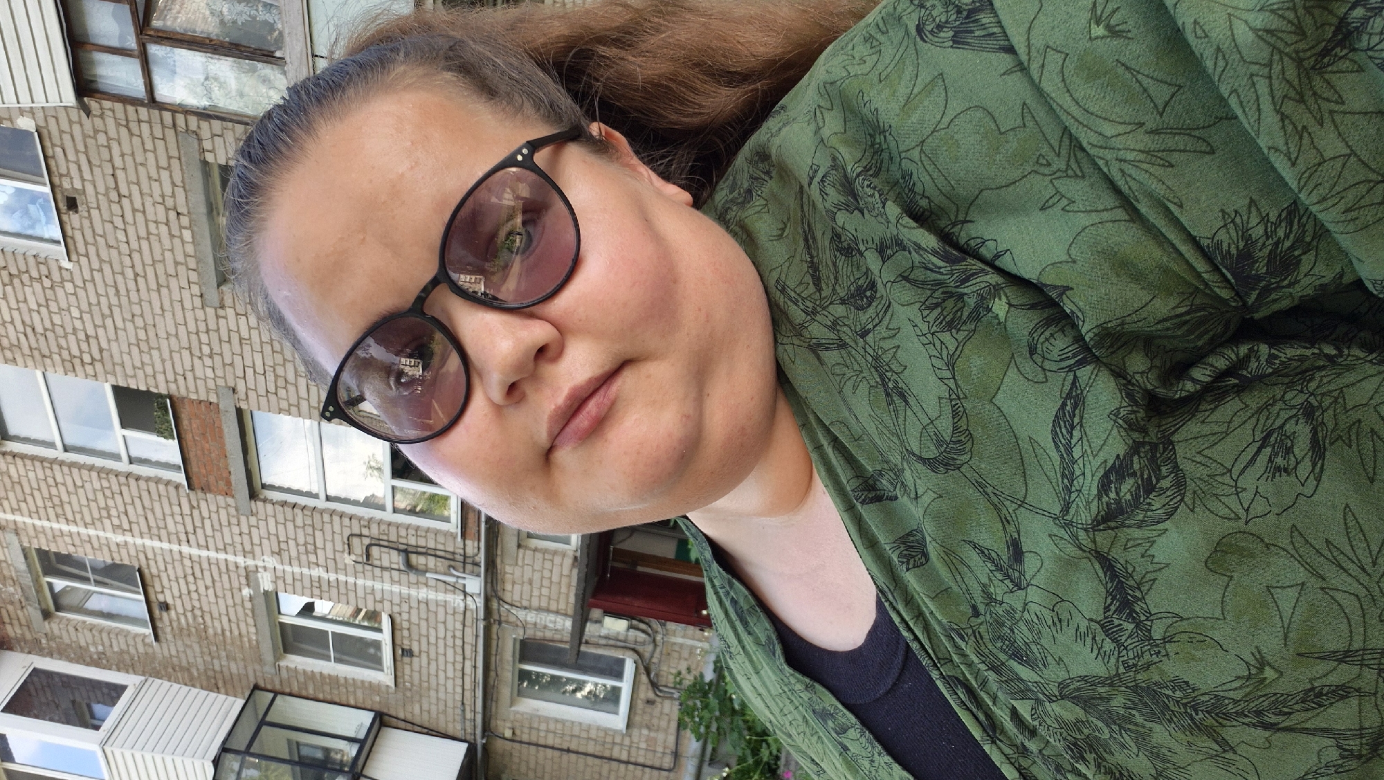 A selfie shows a woman wearing glasses and a green floral print shirt against a brick building. The woman has brown hair and appears to be outdoors. The building has several windows and a balcony. The overall setting suggests an urban or residential area.