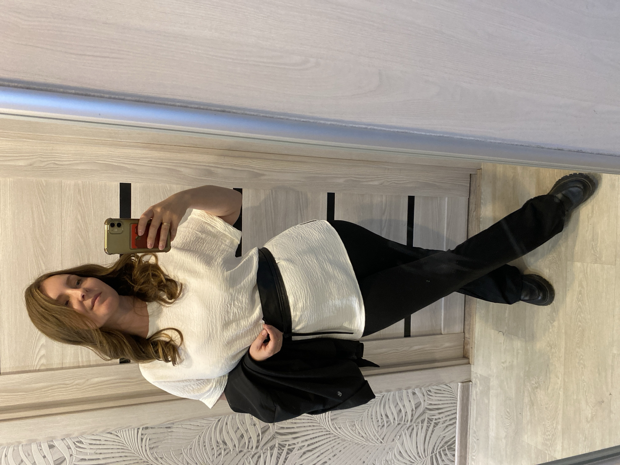 A woman is taking a selfie in a mirror indoors. She is wearing a white, textured top, black pants, and black shoes. She has long, light brown hair. She is holding a gold-colored phone in her right hand. She also has a black jacket draped over her right arm. The background includes a light-colored patterned wall covering, a light-colored door with dark vertical accents, and light-colored flooring. The location information, "Noviy, Russia," cannot be verified from the image.