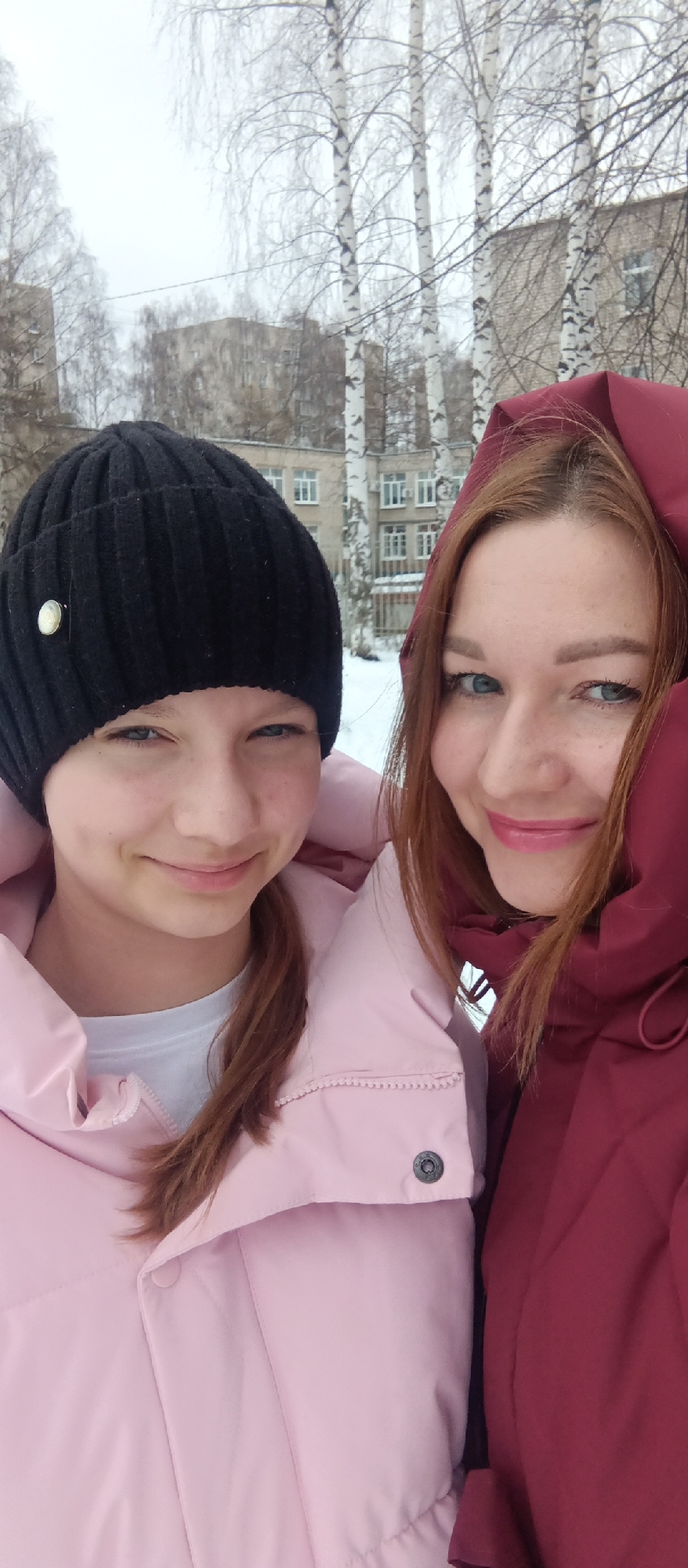 Two women pose closely for a selfie outdoors in an urban environment. The woman on the left wears a black beanie with a silver button and a light pink puffer jacket. She has fair skin and light-colored eyes. Her reddish-brown hair is pulled back into a ponytail. The woman on the right is wearing a red hooded jacket or coat. She has fair skin, reddish hair, and similarly light-colored eyes with freckles. In the background, there are leafless birch trees with white bark, and several buildings, possibly apartment complexes, are visible. The sky is overcast, suggesting a cold or winter season. There appears to be snow on the ground.