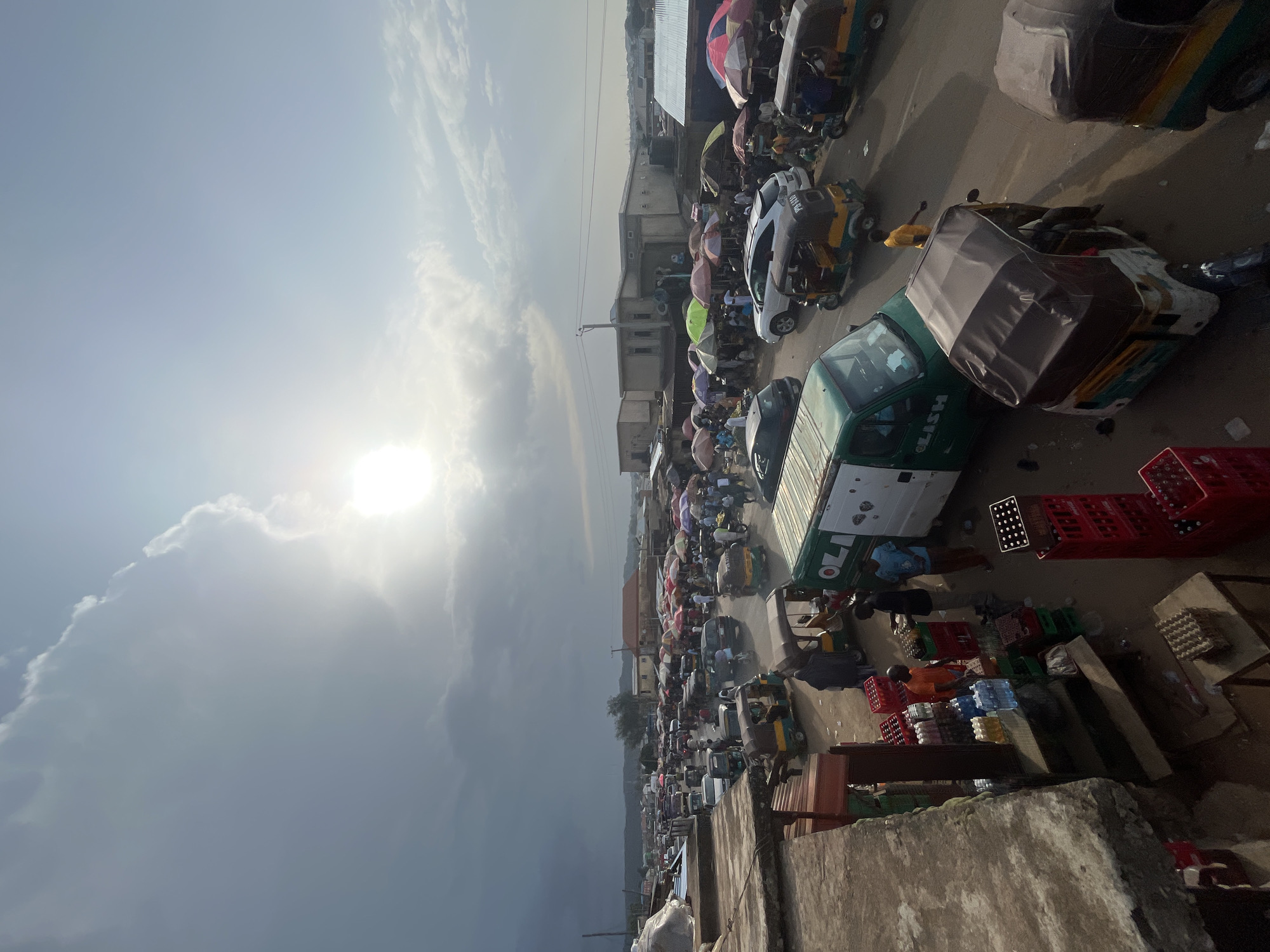 This is a high-angle, wide shot of a busy open-air market in Rafin Par, Nigeria. The sun is bright and partially obscured by clouds, casting long shadows. The market is filled with a variety of vehicles, including numerous three-wheeled auto-rickshaws (Keke Napep) and a few cars and vans. People are walking among stalls, some of which are covered by colorful umbrellas, suggesting a vibrant commercial area. Goods are displayed on the ground and on makeshift tables, including crates of beverages and what appear to be eggs. The overall impression is one of bustling activity in a developing urban environment.