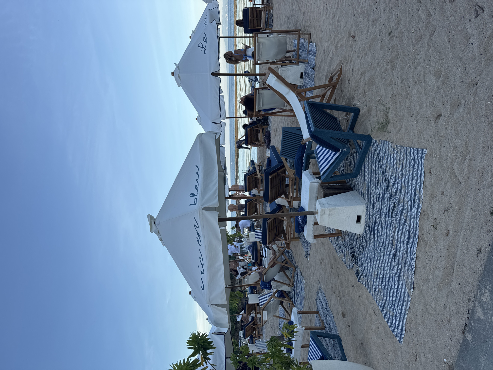 The image depicts a beach scene in Kuta Selatan, Indonesia. Several beach umbrellas, labeled "vie en bleu", are visible, shading groups of lounge chairs arranged on patterned beach mats. People are seated in the chairs, relaxing and engaging in various activities. The beach slopes toward the ocean, visible in the background. The sky is clear and blue. Vegetation is seen along the lower edge of the image. The overall atmosphere is relaxed and leisurely.