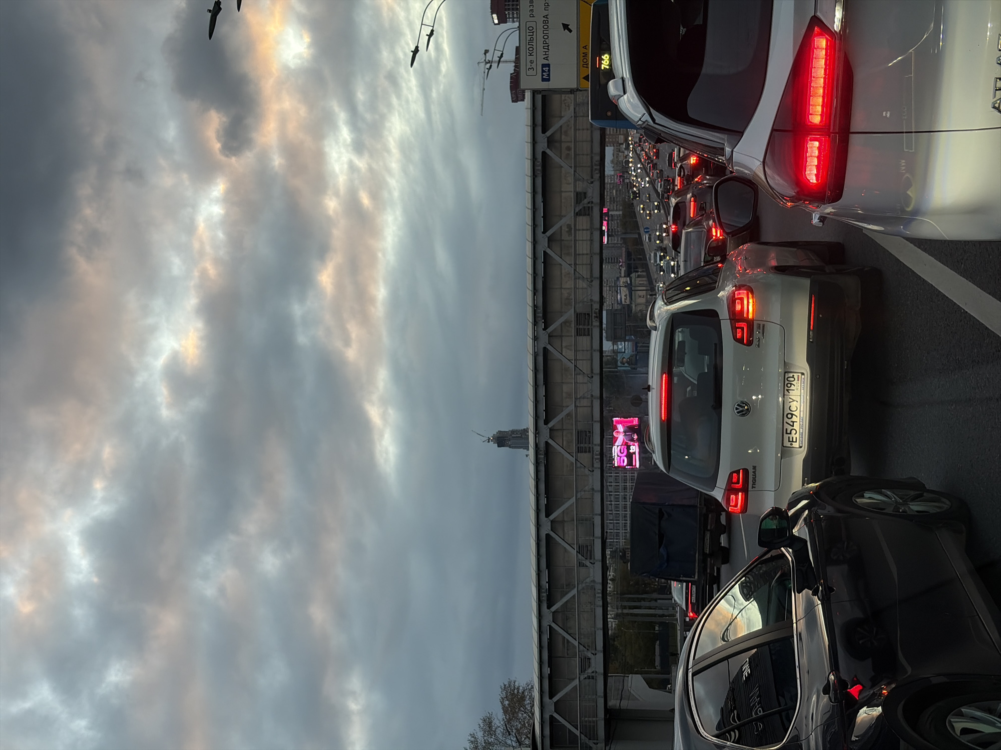 The image depicts a traffic jam on a multi-lane highway in Moscow, Russia, during overcast conditions with hints of sunset colors in the sky. Several cars are stationary, with their brake lights illuminated. In the foreground, a black car is on the left, and a white Volkswagen Tiguan with a Russian license plate reading "E549CY 190" is in the center. To the right, the rear of a white SUV is visible. An elevated pedestrian bridge spans across the highway. In the background, there is a tall building under construction, a digital billboard displaying "5G," and a traffic sign indicating "M4 ANDROPOVA PR." and "3-E KOL'TSO." The sky is filled with dramatic clouds, with patches of pale orange and yellow light. The overall scene conveys a sense of urban congestion.