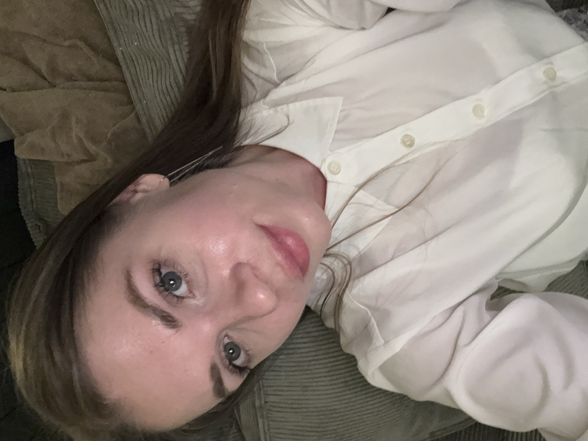 A woman with light skin, brown hair, and blue-gray eyes is lying down. She is wearing a white button-down shirt. The background appears to be a textured, possibly corduroy surface. The location is not discernible from the image.