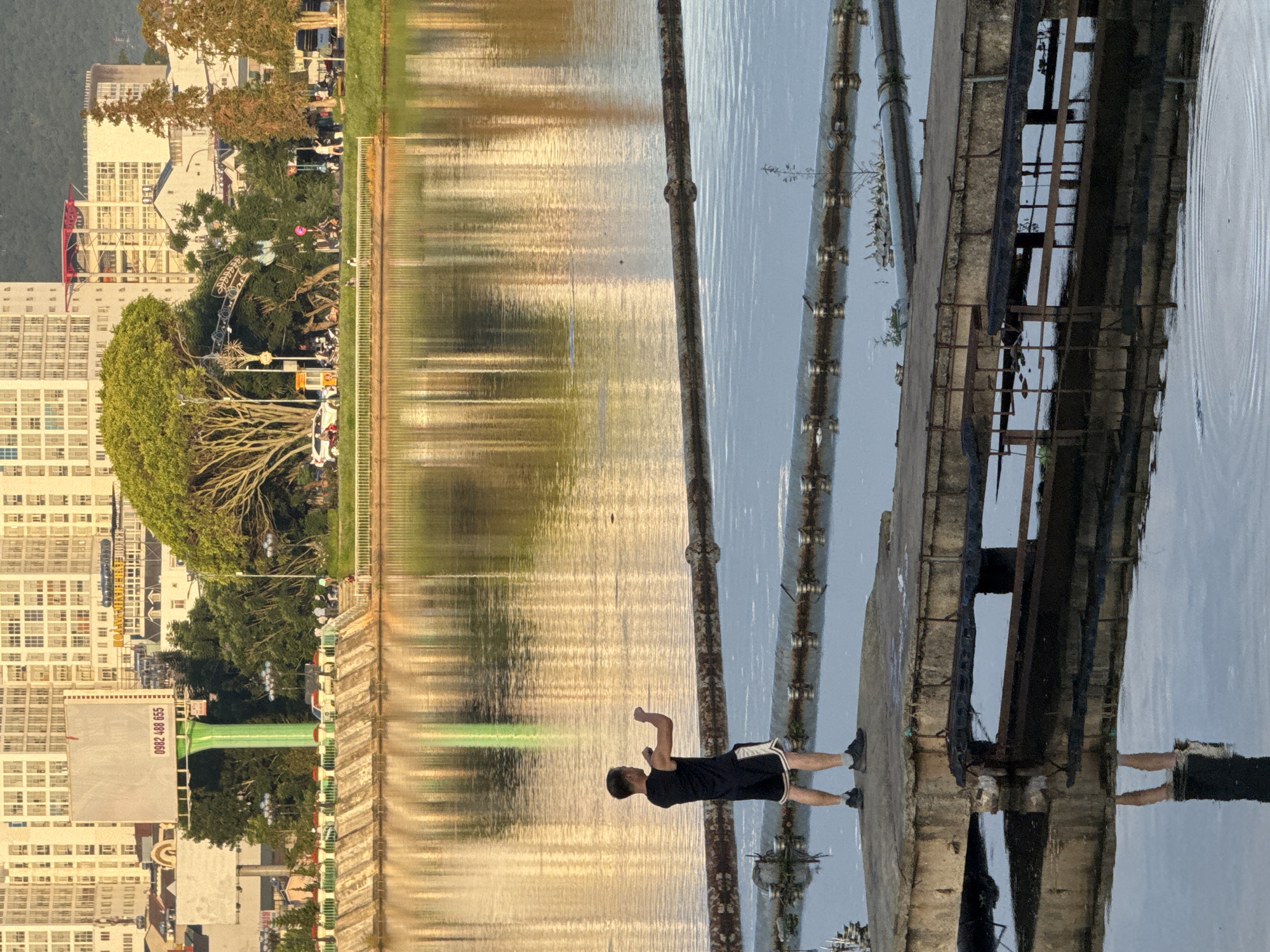 The image depicts a serene outdoor scene in Da Lat, Vietnam, characterized by calm waters reflecting the surrounding urban and natural elements. In the foreground, a young man is captured mid-action, practicing a form of exercise or martial arts on a concrete platform jutting into a body of water. He is dressed in athletic attire, a black tank top and shorts, his posture suggesting focus and physical exertion. The platform he stands on is weathered and appears to be part of an old structure, possibly a pier or a former industrial installation, with visible pipes and conduits running along its length and submerged in the water.
The water itself is a significant element, its surface rippling gently, reflecting the golden hues of the late afternoon or early morning sun. These reflections create an abstract pattern of light and color, partially obscuring the details of the buildings and trees on the opposite bank.
Beyond the water, the cityscape of Da Lat unfolds. In the background, a line of multi-story buildings, typical of urban development, stands tall, their facades reflecting the sunlight. A large billboard with a green support structure is prominently visible, bearing a phone number. Lush green trees and foliage punctuate the urban landscape, creating a contrast between the man-made structures and the natural environment. A road runs along the water's edge, with a few vehicles and pedestrians suggesting a quiet, ordinary day in the city. The overall impression is one of tranquility, with the lone figure engaged in his activity serving as the central focus amidst the expansive, reflective environment.