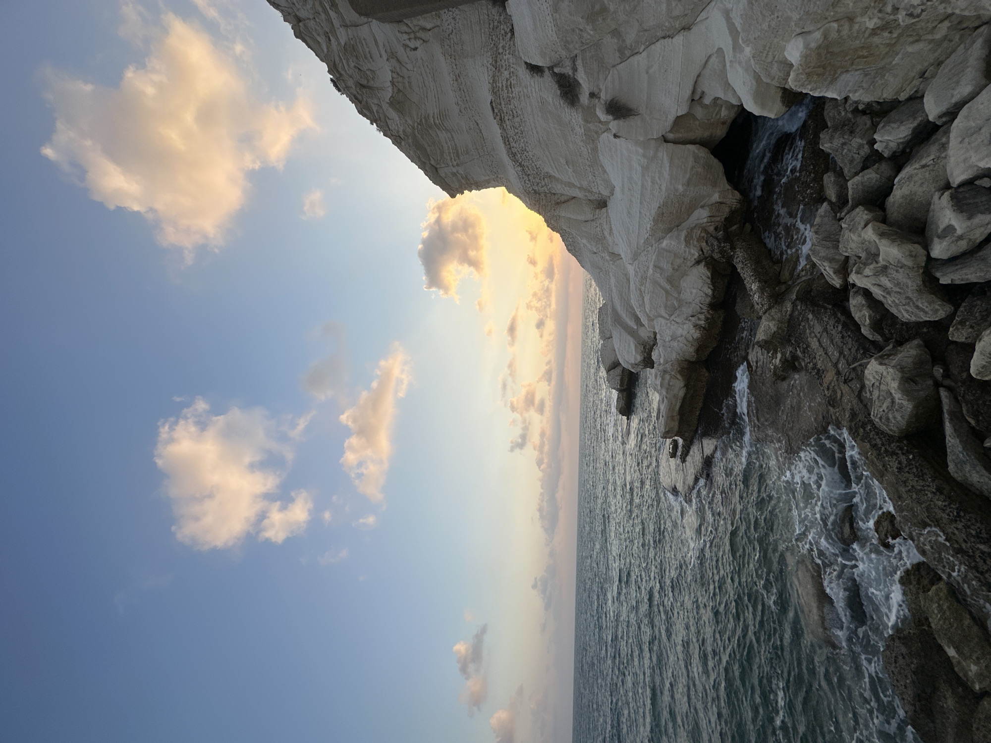 This image depicts a coastal scene at sunset in the city of Mate Asher Regional Council, Israel. The foreground features a rocky shoreline with waves gently crashing against the rocks. The water is a deep blue-green. To the right, a large, white cliff face rises dramatically, showing layered rock formations. The sky above is a soft blue with scattered clouds illuminated by the setting sun, casting a warm orange glow. The horizon line is visible where the sea meets the sky. The scene is devoid of people or any visible structures. The overall impression is one of natural beauty and tranquility.