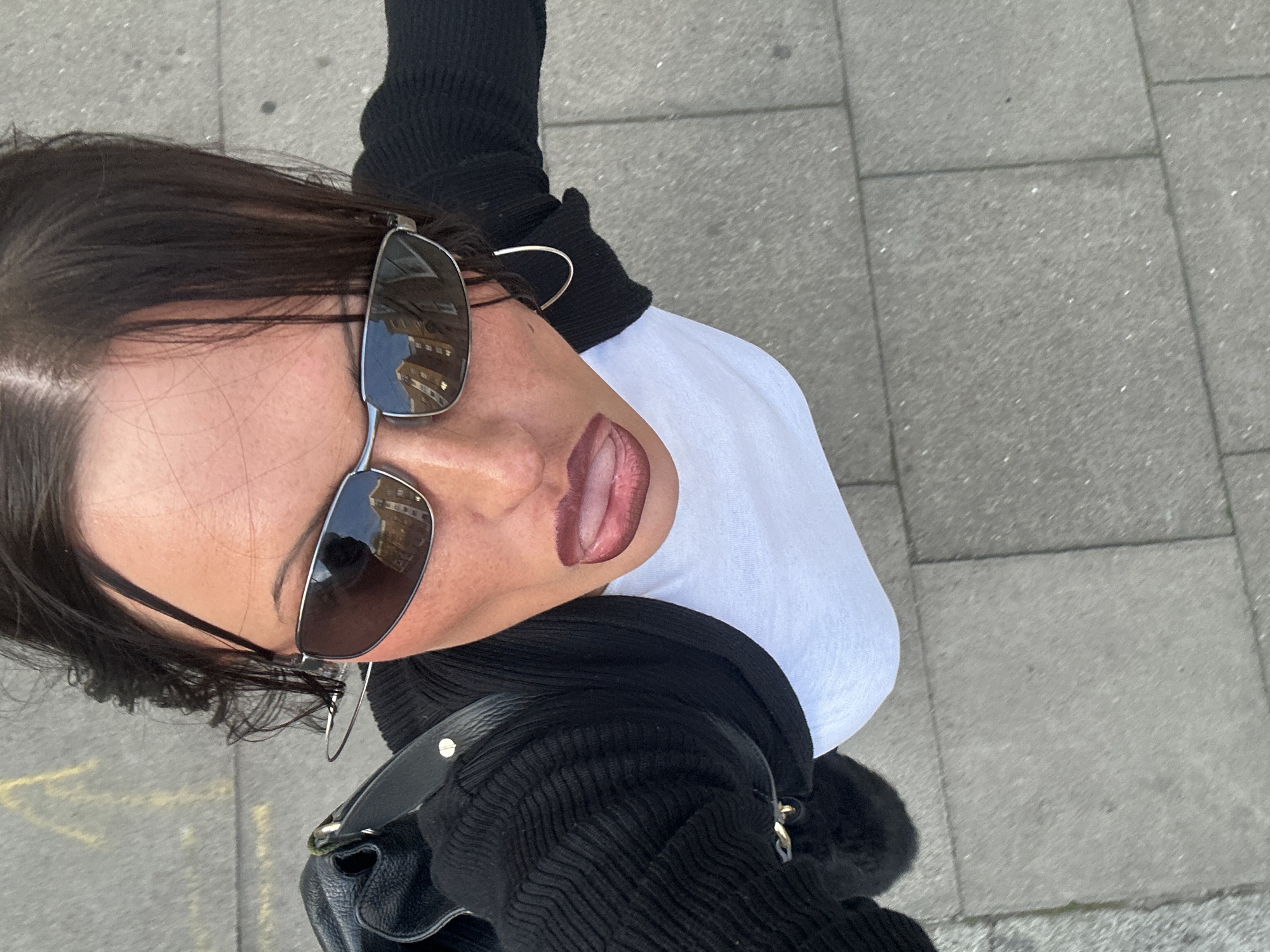 A high-angle, overhead selfie captures a close-up of a person, likely female, from the shoulders up, looking directly into the camera. The subject has dark hair, parted on the side, with some strands falling around the face. They are wearing stylish silver-framed, dark-lensed sunglasses that reflect urban buildings, suggesting a city environment under clear skies. Beneath the sunglasses, the upper part of the nose is visible. The mouth is slightly open with the tongue extended, revealing dark, possibly brown or plum-colored lipstick on the lips.
The person is dressed in a black ribbed long-sleeved top or cardigan, worn over a plain white t-shirt, parts of which are visible on the right side of the frame. A black leather-like bag with a visible gold-colored clasp or hardware is slung over their shoulder, partially seen in the lower left.
The background consists of grey concrete paving slabs that form a sidewalk or urban pathway. The bright, even lighting indicates it is daytime. The overall impression is a casual, confident, and perhaps playful moment captured outdoors in an urban setting, consistent with London, United Kingdom. No visible text is present in the image.