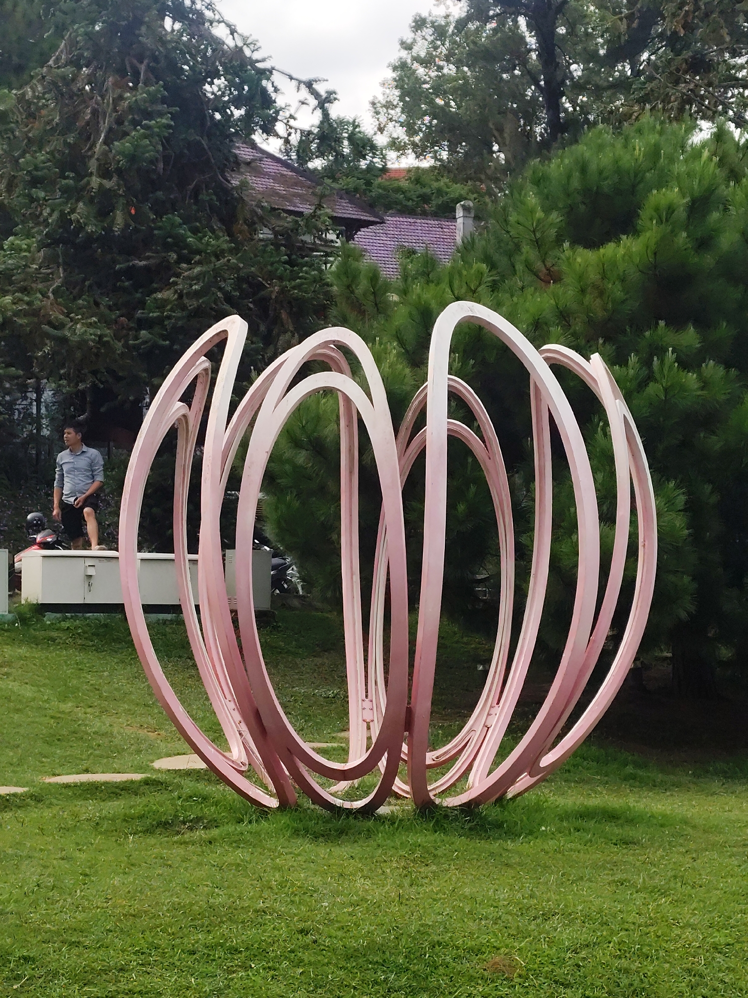 In Da Lat, Vietnam, a large, pink, circular sculpture stands on a green grassy lawn in front of a backdrop of dense evergreen trees and a building with a purple roof. A young man in a blue collared shirt and black shorts is standing on top of a white rectangular structure to the left of the sculpture, looking to his right. A motorcycle is parked to the left of the white structure. The sculpture is composed of numerous interconnected rings that form a spherical shape. The scene is outdoors and appears to be a park or garden setting.
